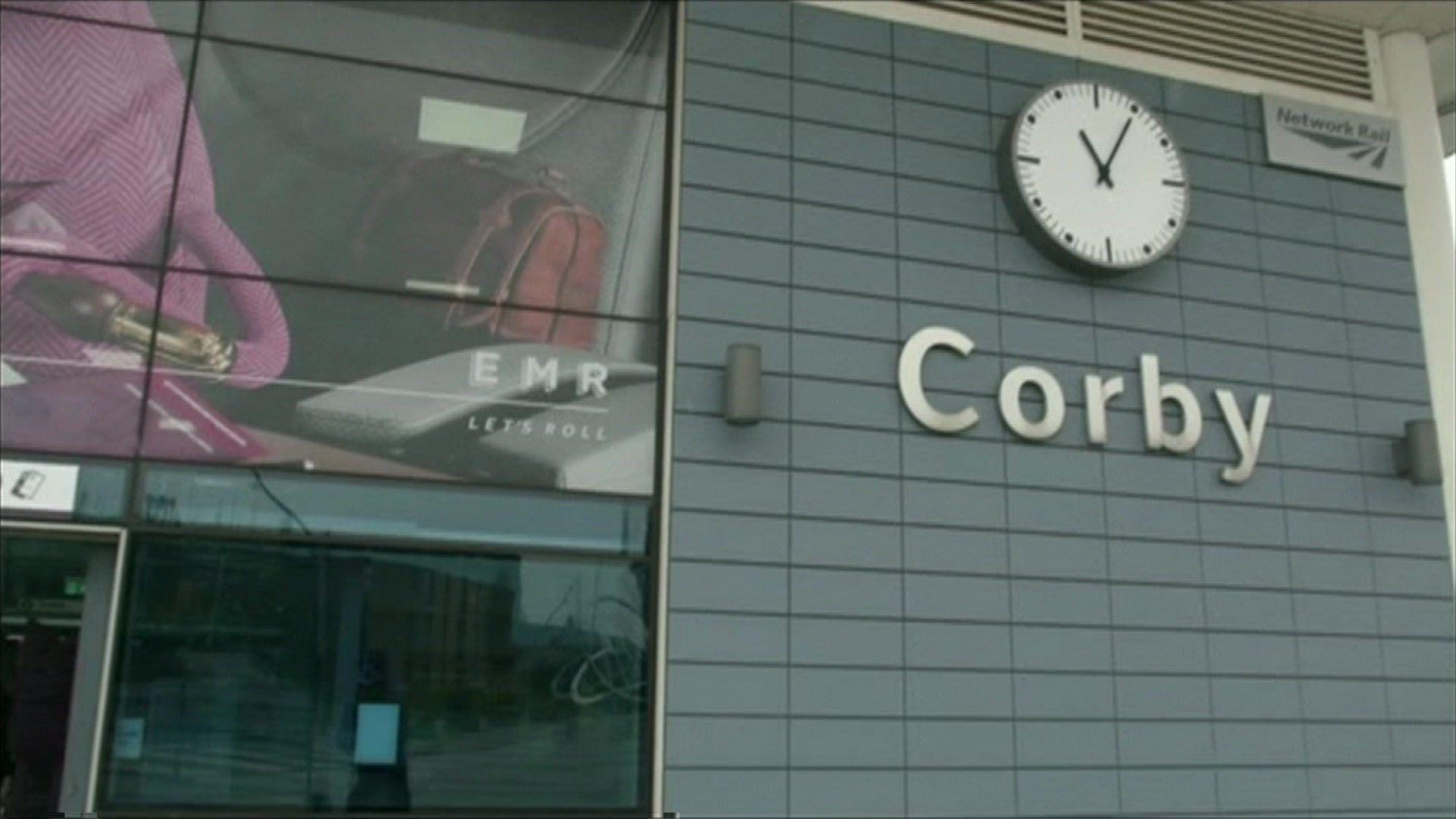 Corby Railway Station's frontage has a large sign with the town's name and a clock above that. Adverts can be seen on the left of the image