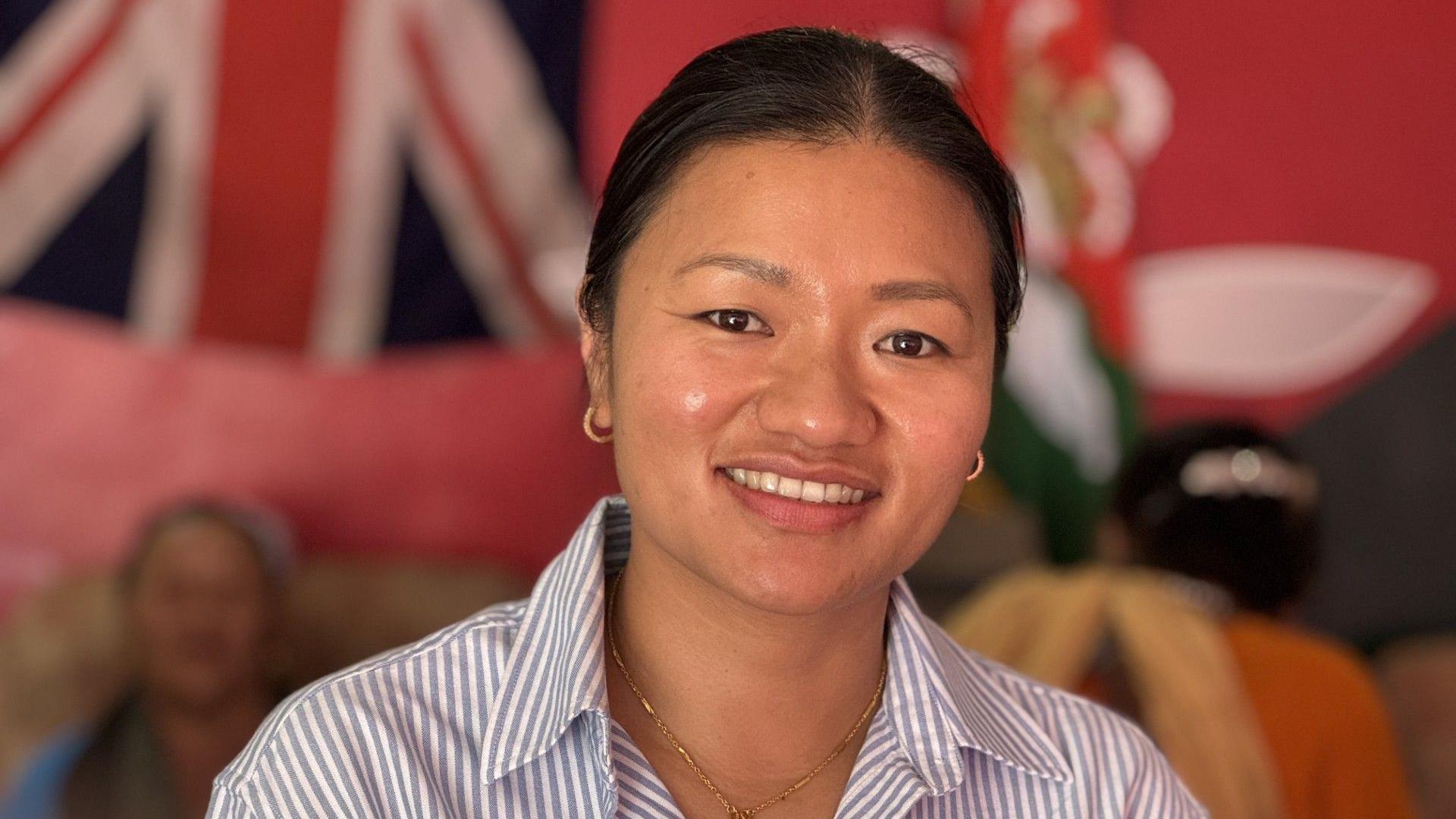 A smiling Pratikshya Gurung is standing in front of a Union Jack flag with a couple of older Gurka women sitting behind her. She's wearing an open necked blue and white striped shirt, revealing a small necklass with a cross on it.