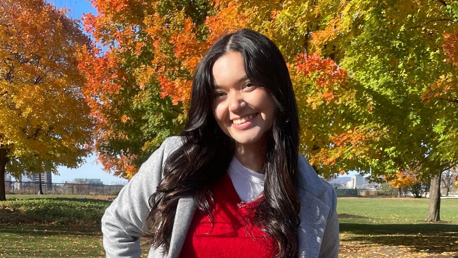 Elizabeth Jerome stands outdoors surrounded by trees in a red jumper and grey coat. She has long dark hair and is smiling at the camera.