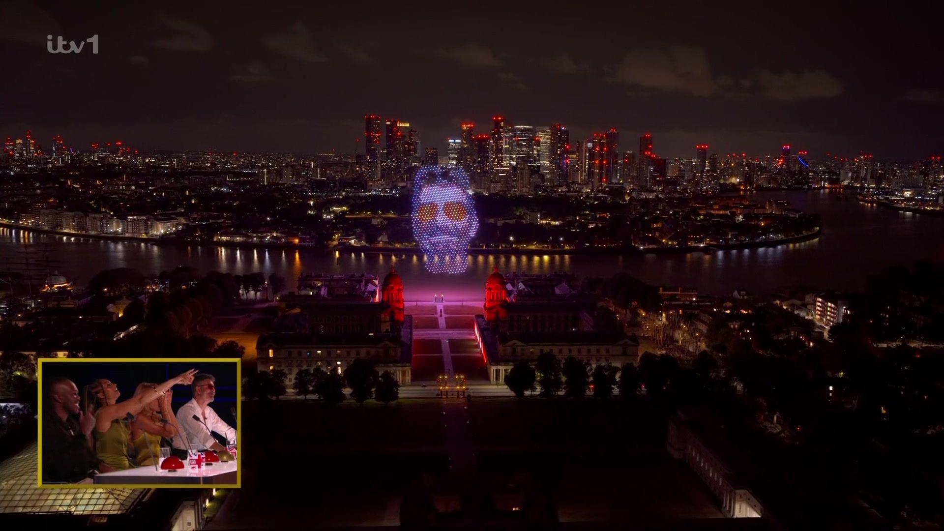 A drone show over London's skyline. Simon Cowell's face is projected in lights. In a reactioj shot from the studio we see the judges smiling and pointing