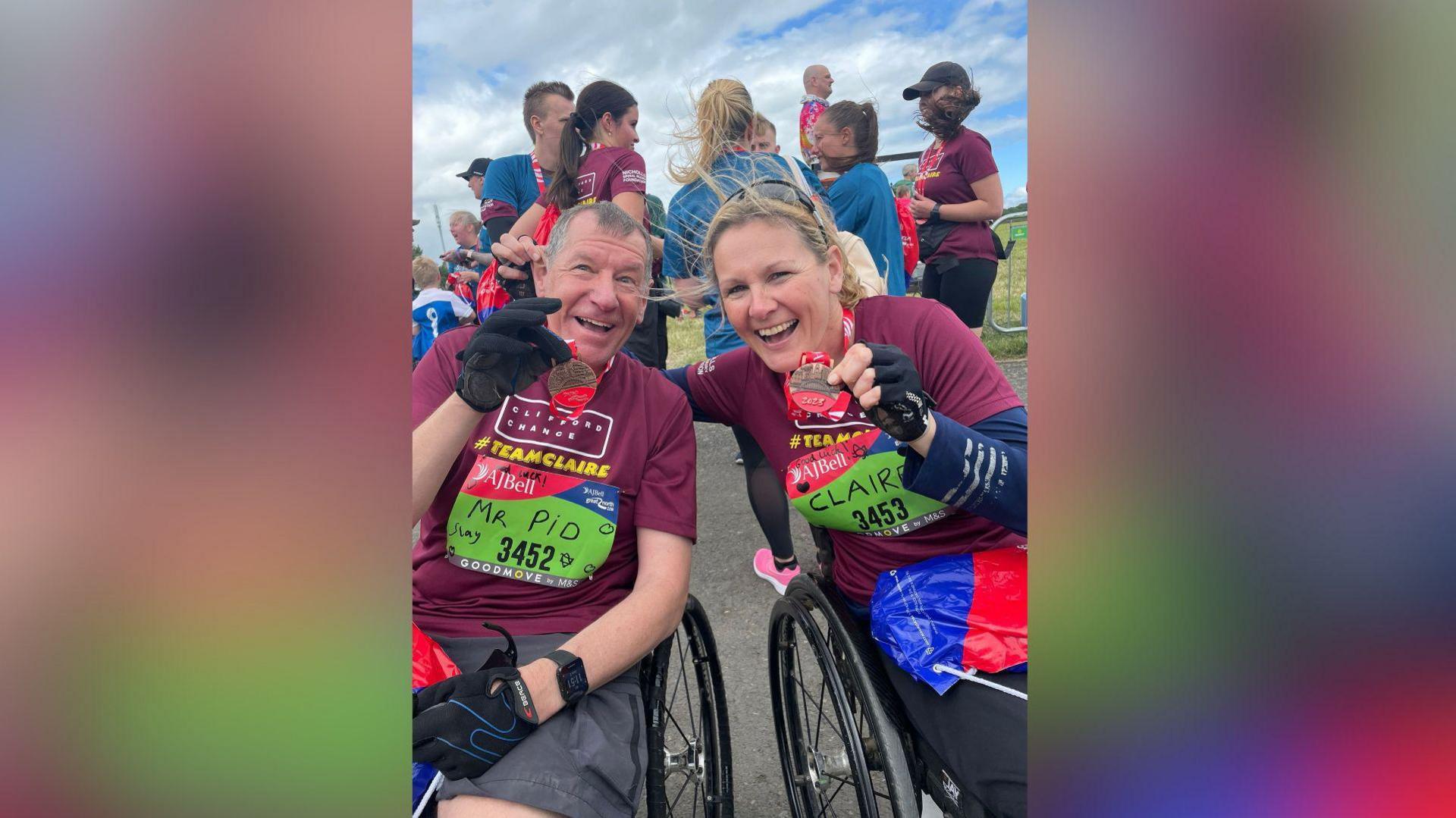 Stuart Hall and Claire Lomas in wheelchairs, both wearing burgundy tops, with medals around their necks after completing a challenge