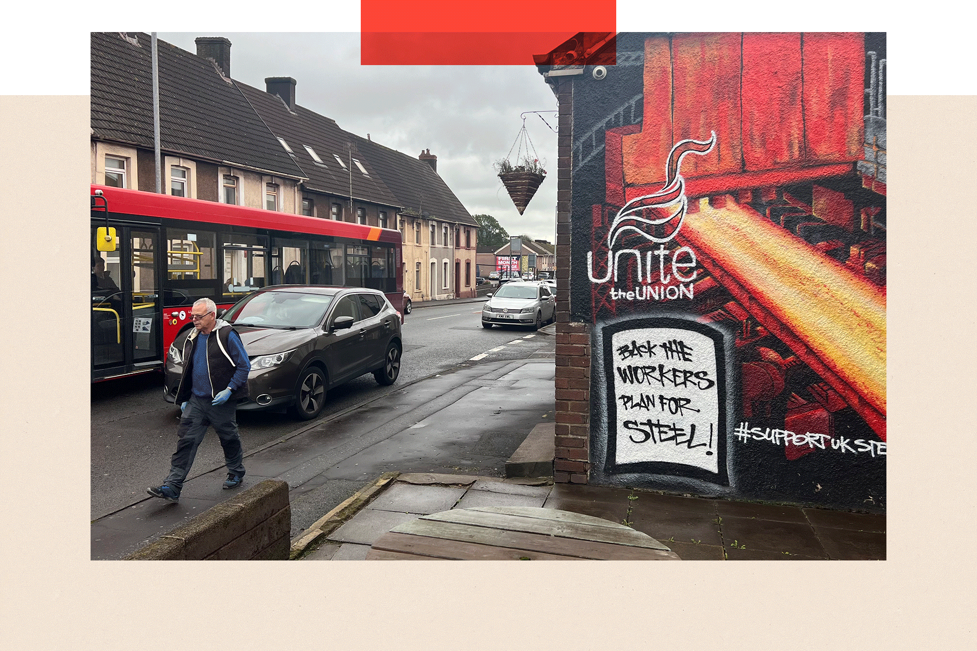 Terraced houses on a street with a red bus and man passing by and a Unite union mural on a wall saying 'back the workers plan for steel!'