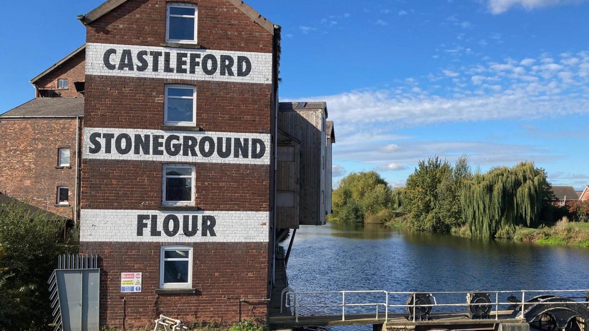 A brick building bearing the words 'Castleford Stoneground Flour' stands next to a river. On the other side of the river are several trees.