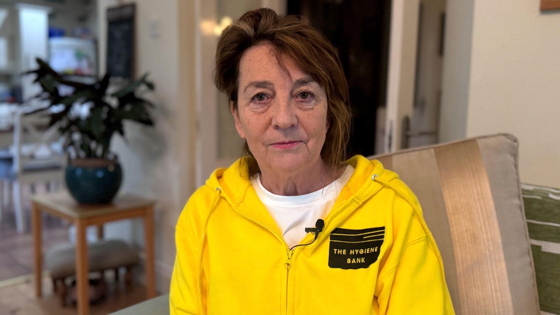 Eileen Brown with short brown hair and is pictured staring at the camera as she wears a yellow zip-up jumper with 'The Hygiene Bank' written on it. She is sitting on a sofa and there is a table with a plant pot on it in the background.