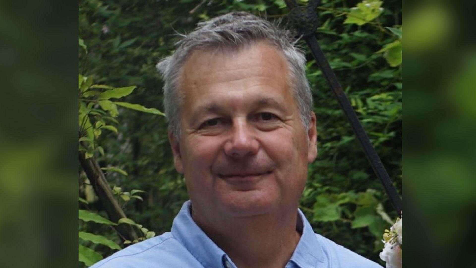 Ian Gooden, with short grey hair, is wearing an open-necked light-blue shirt. Behind him are lush green trees and plants. 