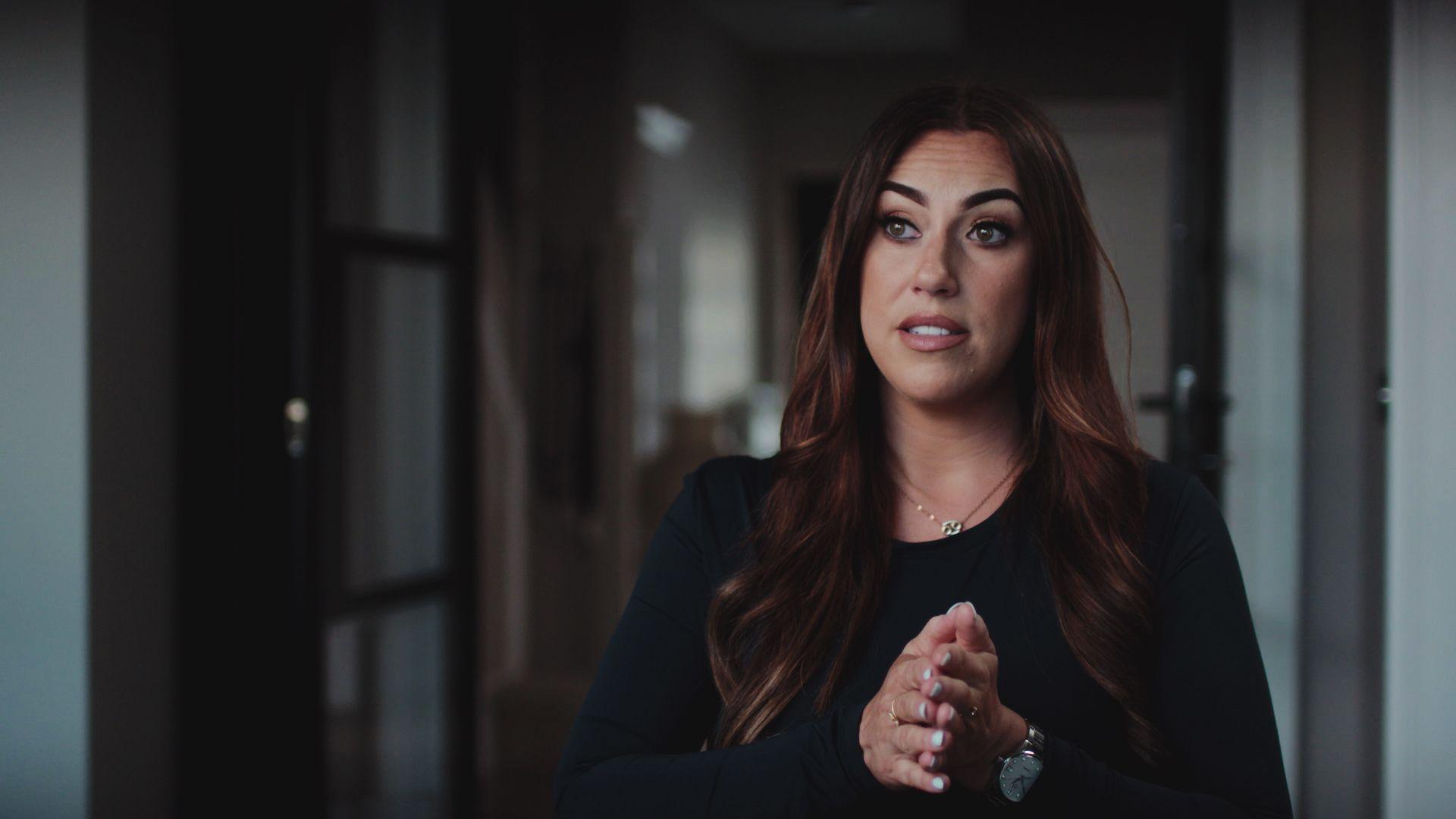 Aimee Tennee sitting in a room with white walls an a dark wood door. She has long brown hair and is wearing a black top.