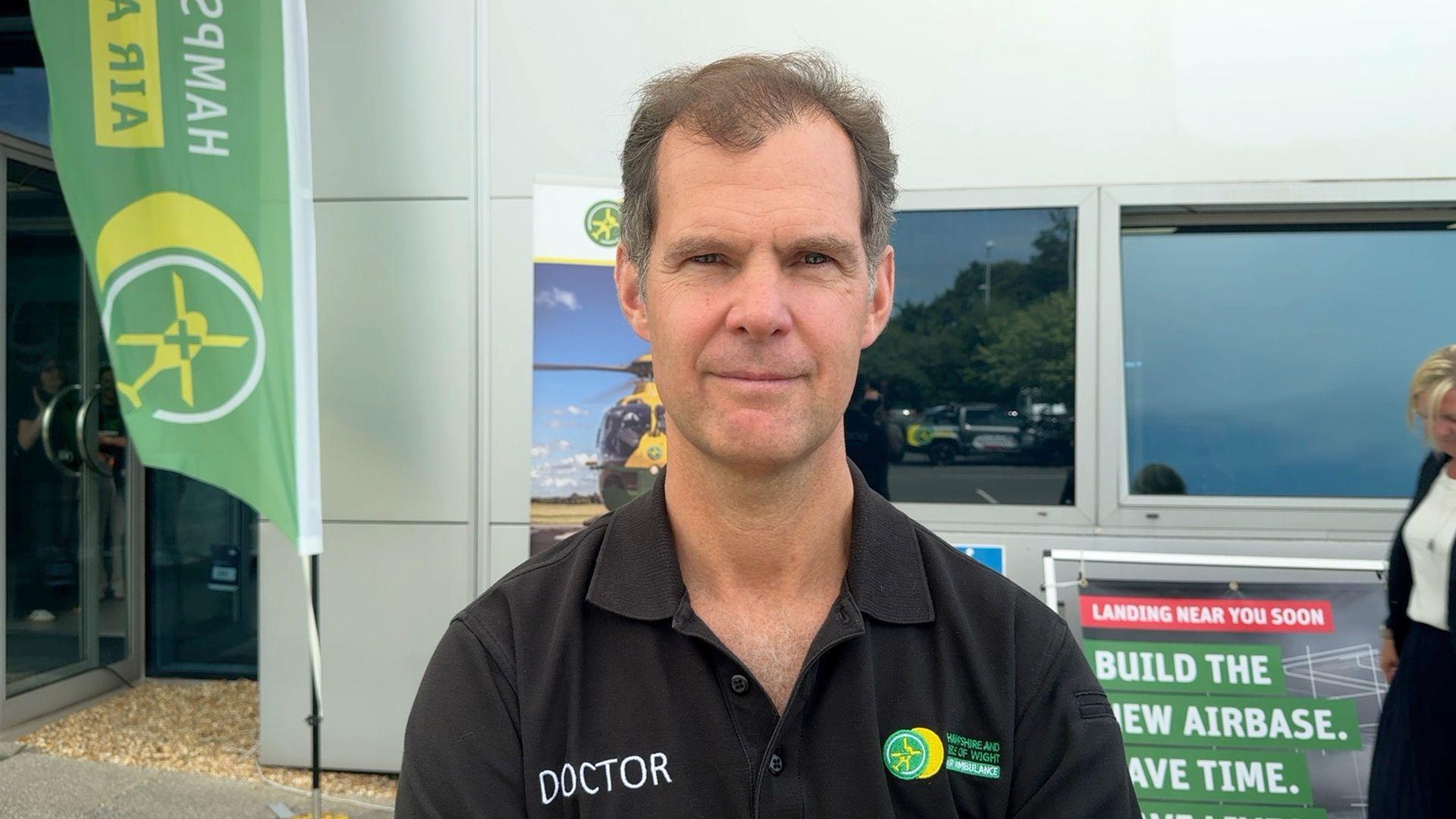 A man is looking towards the camera wearing a black polo shirt. The words 'Doctor' and 'Hampshire and Isle of Wight Air Ambulance are printed on the front. He has balding black hair.