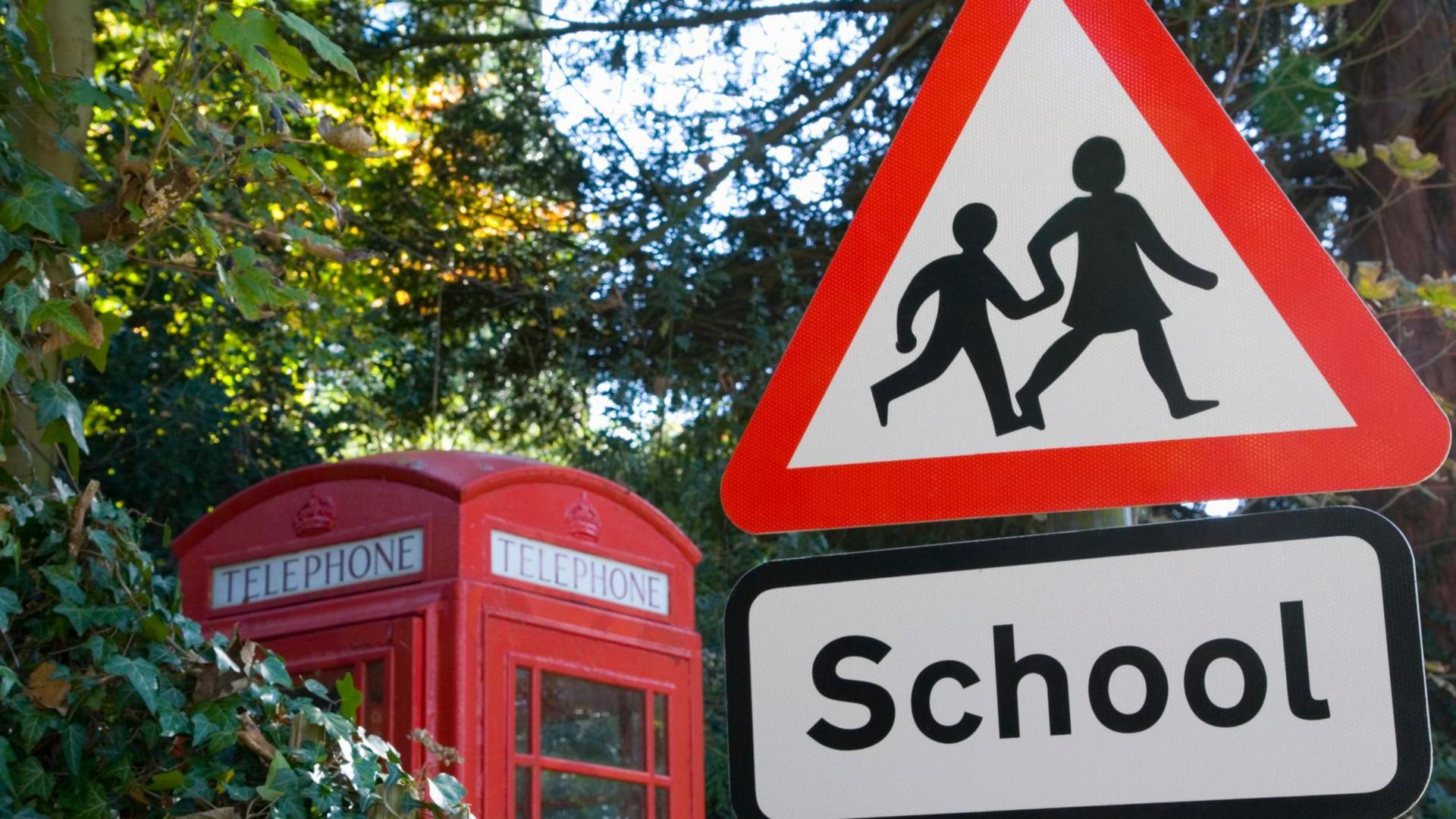 A school crossing sign in red and white with an old-fashioned red public telephone box behind it