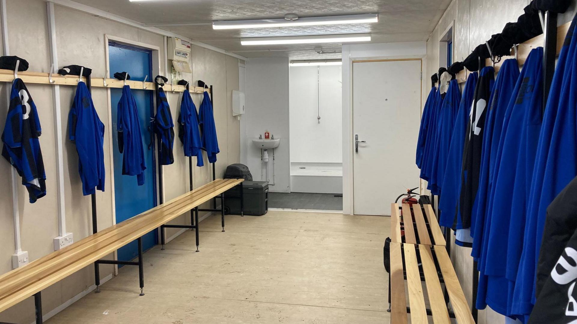 A picture showing the new changing rooms. There are blue football kits hung up on the wall with wooden benches underneath.