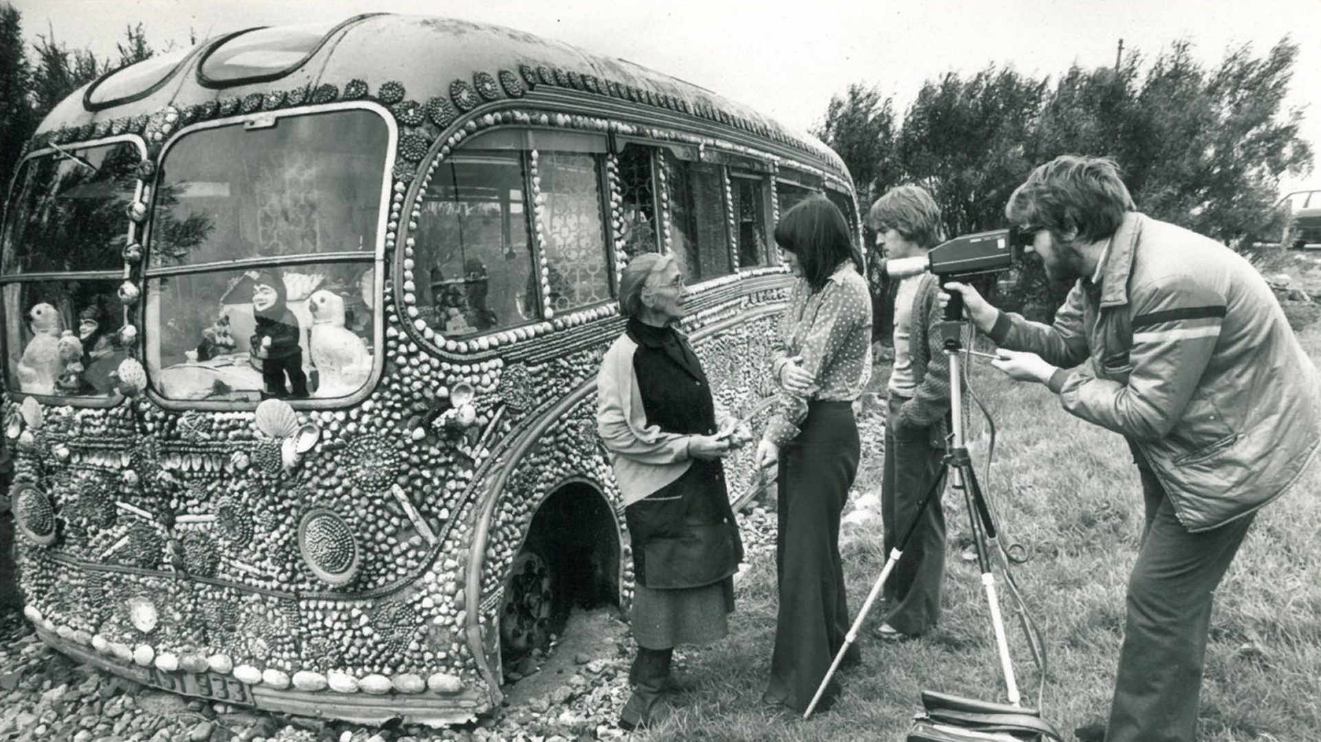 A bearded man in an anorak is looking through the viewfinder of a video camera at an elderly woman who is being interviewed on tape by a younger woman. A young man is watching on. They are standing beside a dilapidated bus which is covered in sea shells and has dolls in the windows.