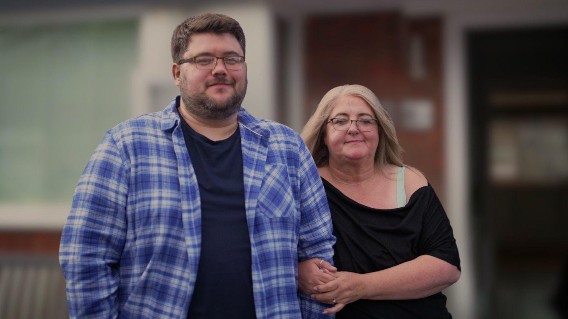 Liam and his mother Melissa stand outside a house, which is blurred in the background. They are both smiling, and Melissa has her arm linked around Liam's affectionately. 