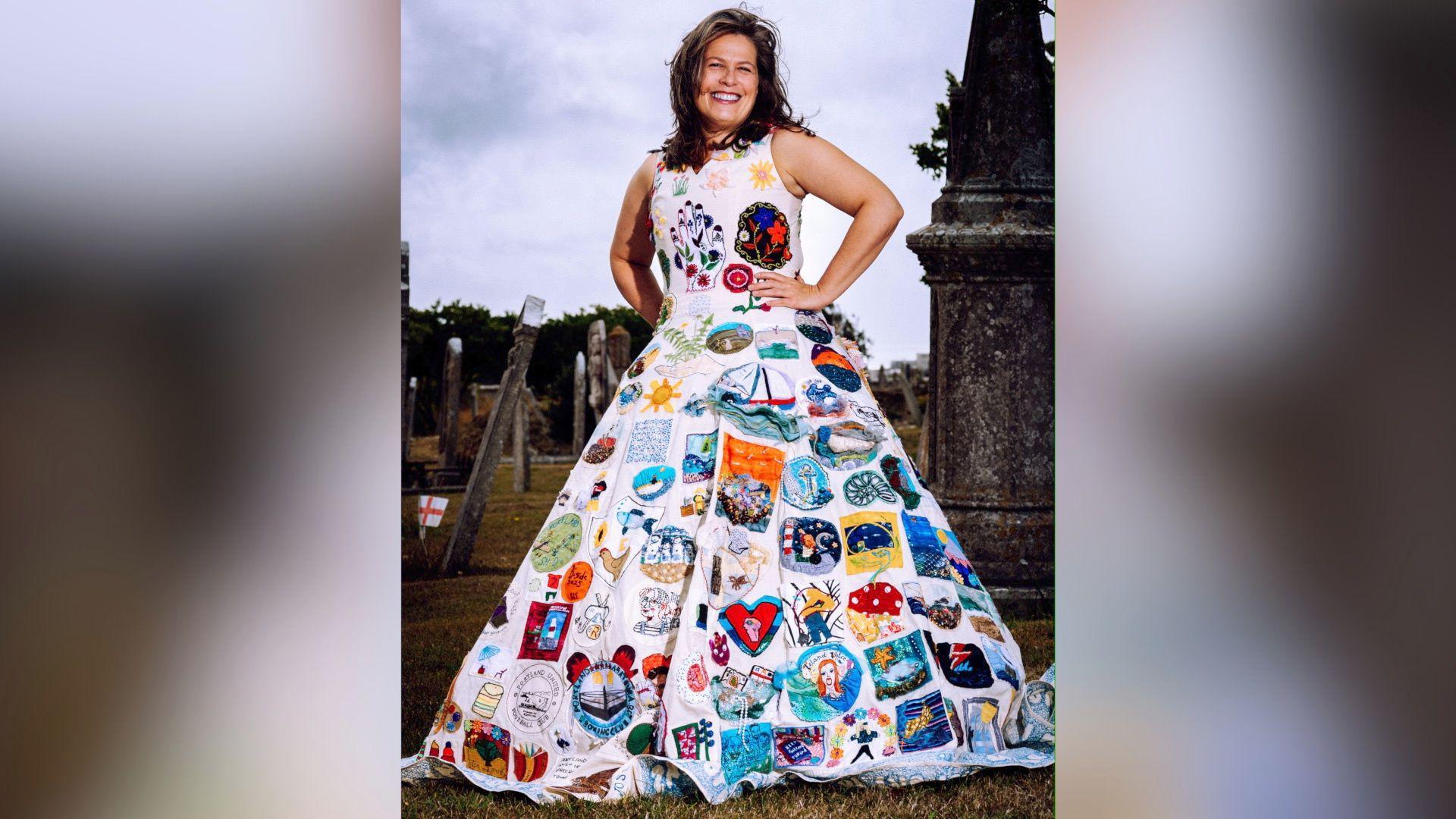 Annerose Walker is standing outside in a graveyard. She has her hands on her hips and is grinning widely at the camera. Her dark brown her is windswept around her face. She is modelling the Portland Dress.