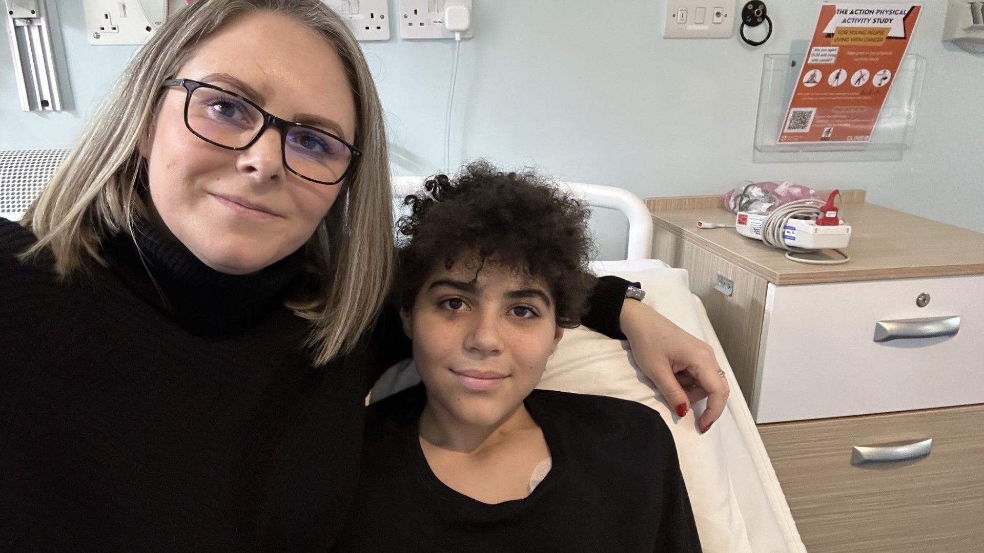 Helena next to Zak on a hospital bed. Helena has her arm around Zak, they are both looking at the camera and smiling.