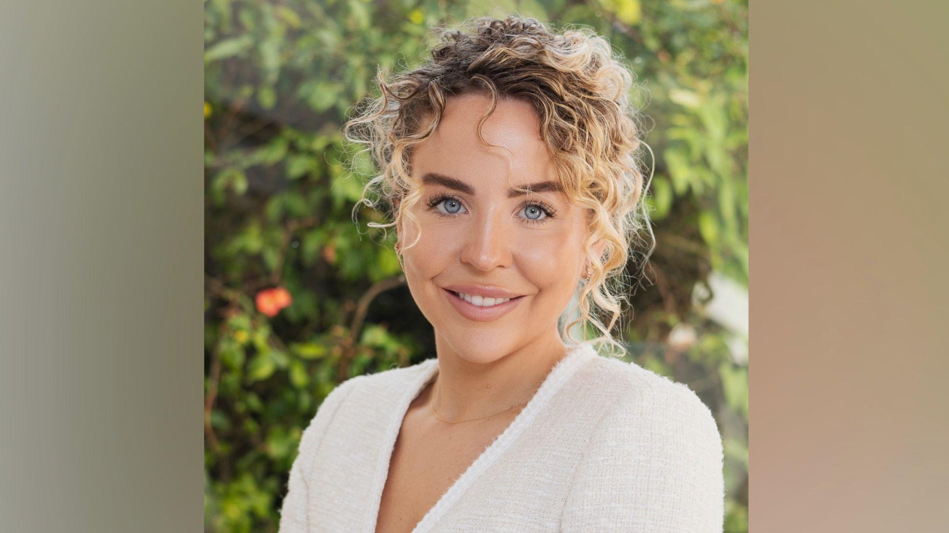 Lydia Bright wears a white top with lace trim. She has blonde curly hair which is tied back. She is sitting in front of a blurred background of green foliage.