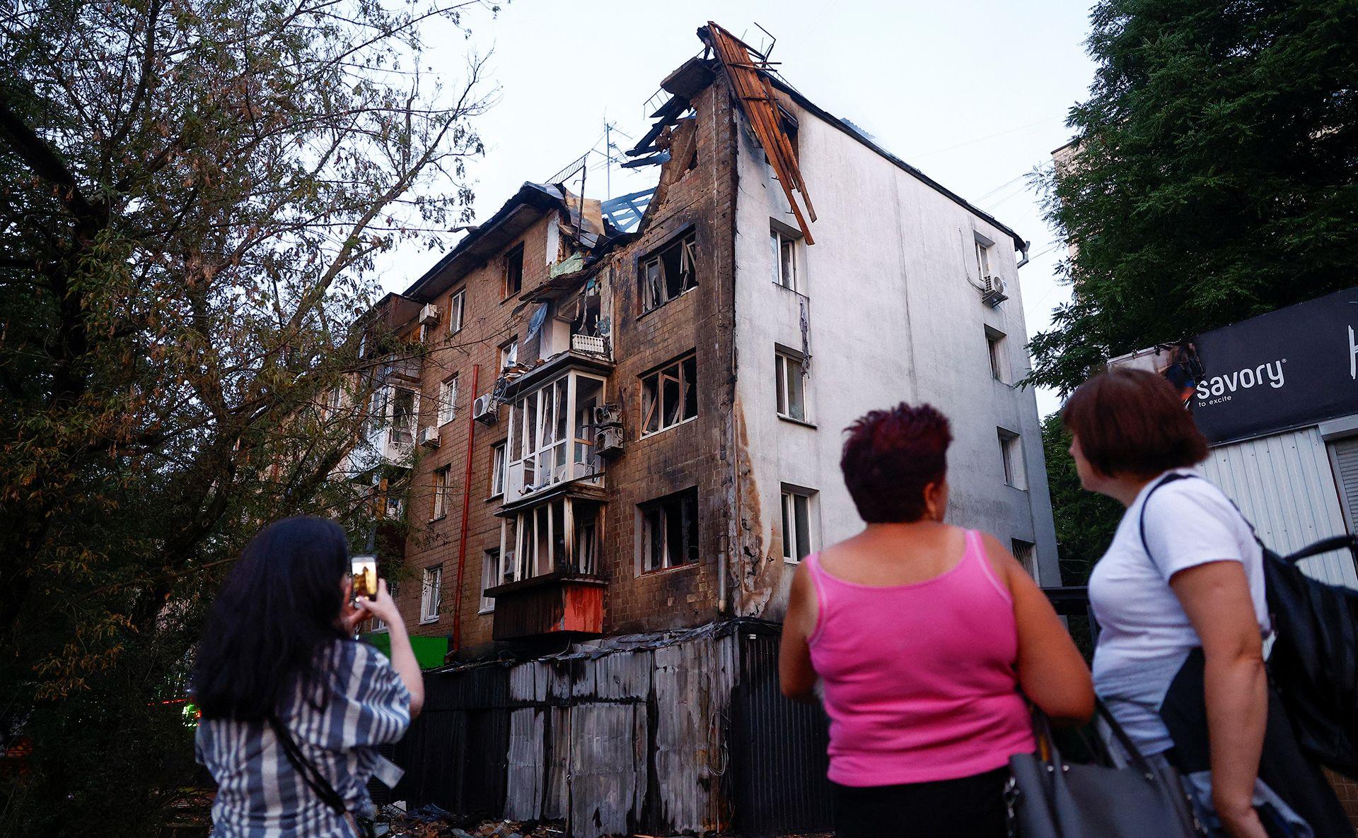 People stand outside looking at a residential building in Kyiv damaged in a Russian drone attack, with one taking a photo on their phone. The building is visibly damaged with a large hole on the top floor and trees line the street, in Kyiv on 10 July.