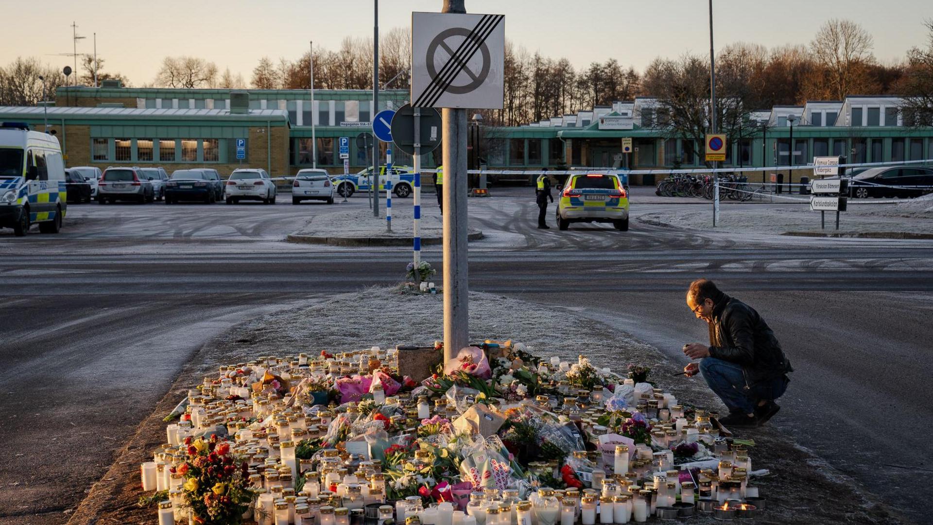 A cluster of candles and flowers lies on the frosty ground as a man crouches in front of the scene