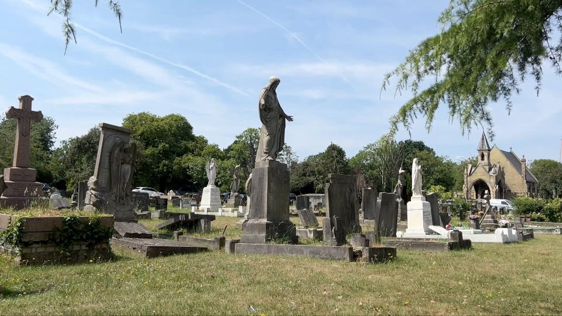 A cemetery with a variety of tombstones, stone crosses, and statues, including religious figures. In the background stands a small chapel or church with a pointed roof, surrounded by trees under a clear sky.