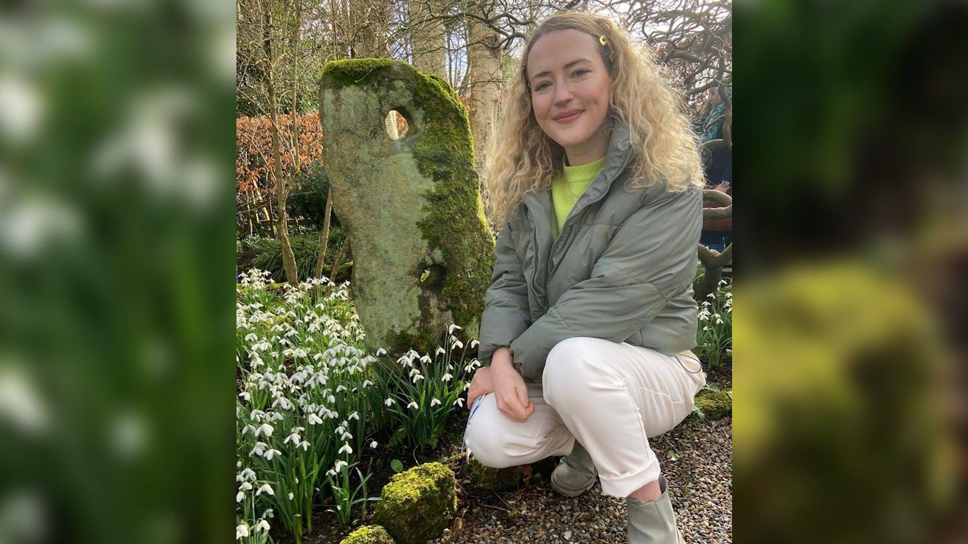 A smiling woman with long blonde hair, wearing white trousers and a green jacket. She is kneeling down by a small decorative standing stone. The stone is surrounded by snow drops in bloom, with trees in the background.