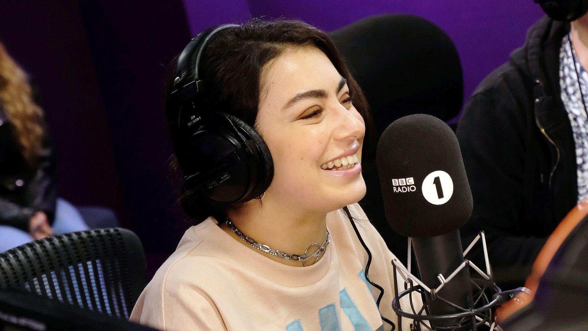 Charli XCX has dark hair tied up. She is wearing headphones and smiling in front of a 91ȱ Radio 1 microphone. She is wearing a silver chain and a peach tshirt. 