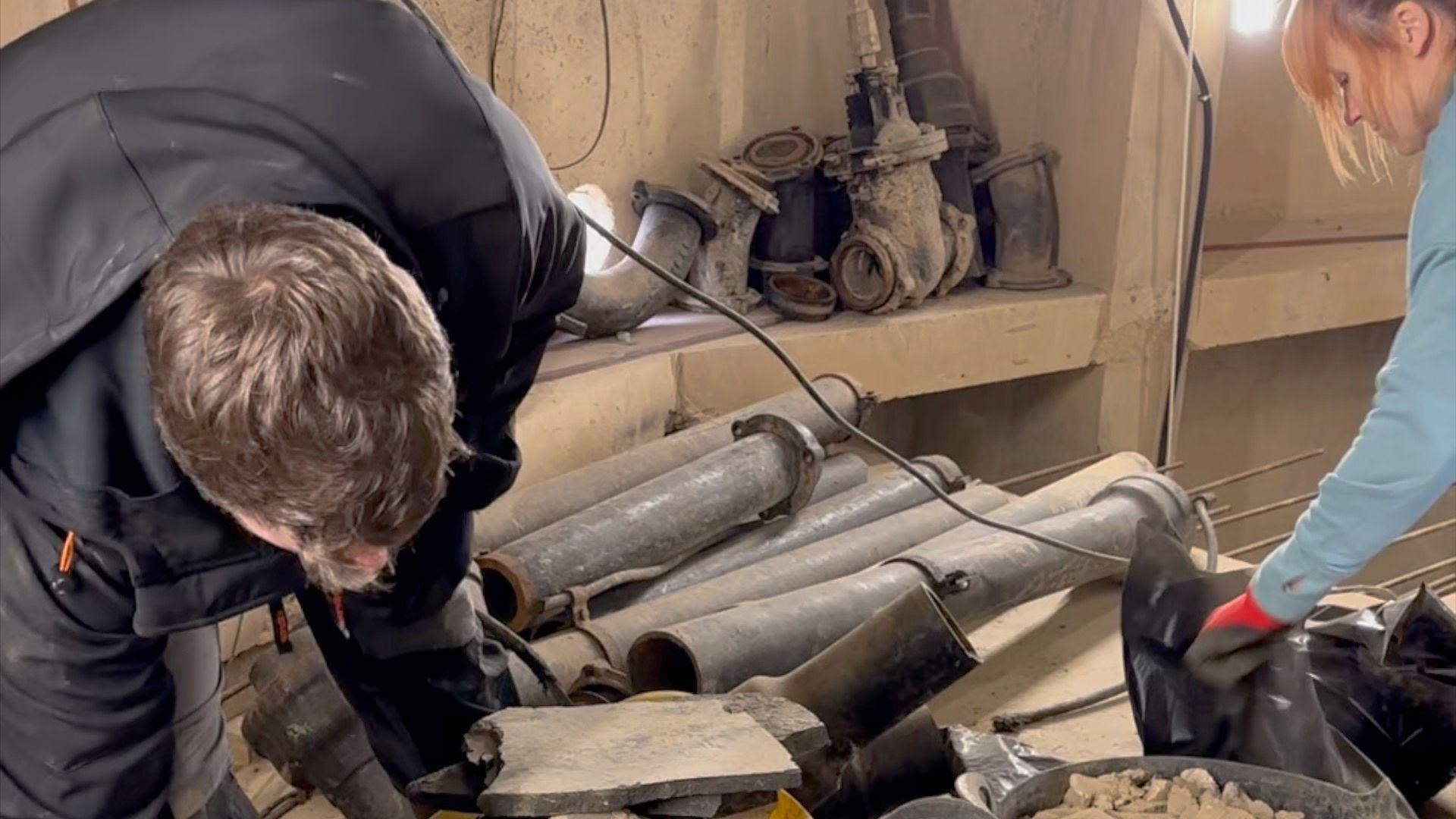 Adam, on the left, in a black jacket, clearing a room filled with old metal pipes inside the water tower. Tassy, on the right, is also working in the room. 