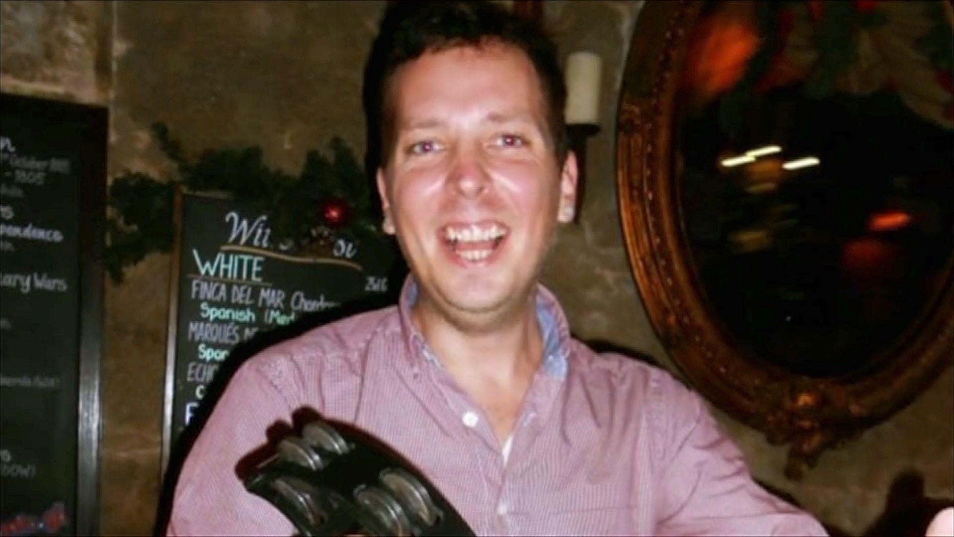 A man is looking at the camera wearing a red shirt. He is smiling. He appears to be a in a dark pub.