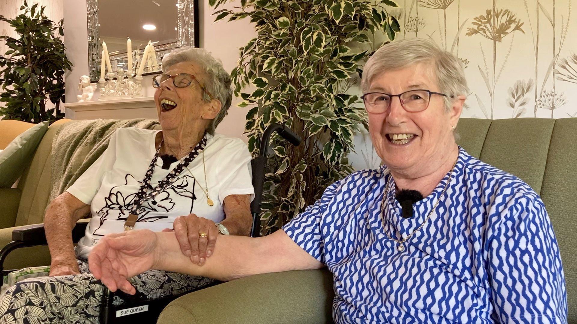 Two older ladies, one wearing a white T-shirt and the other in a blue and white top, share a laugh together. One has her head thrown back laughing and is clasping her friend's arm.