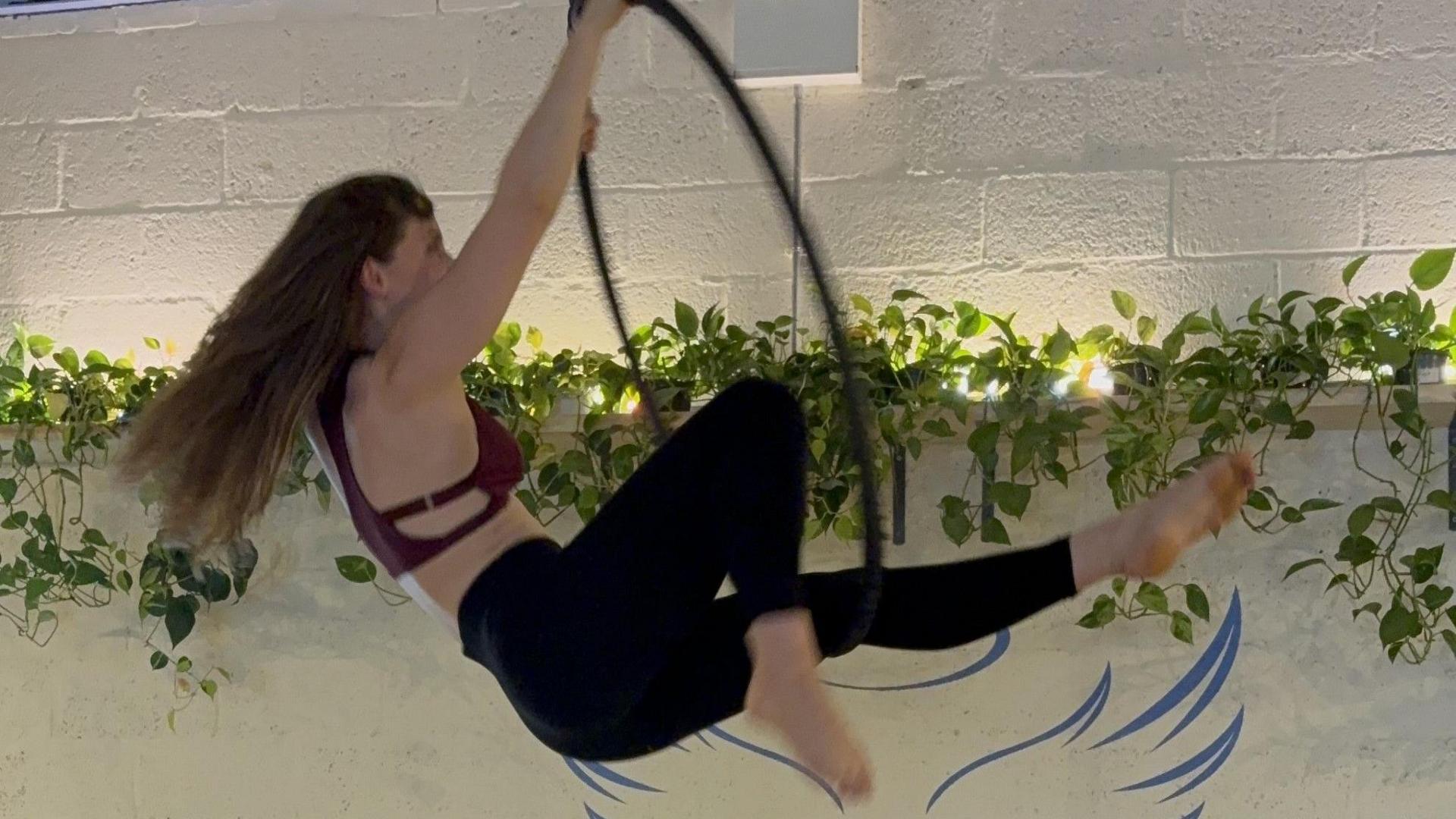 Giselle Pearson performing at the aerial dance studio. She is using a hoop and has one leg outstretched and the other bent, and her long brown hair is streaming behind her as she moves.