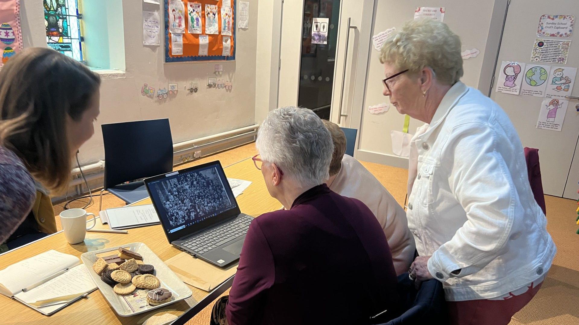 Ella, Catherine and Georgina with their back to the camera, looking at a black computer with a screen showing an old picture. A variety of biscuits are to the far left of the picture.  