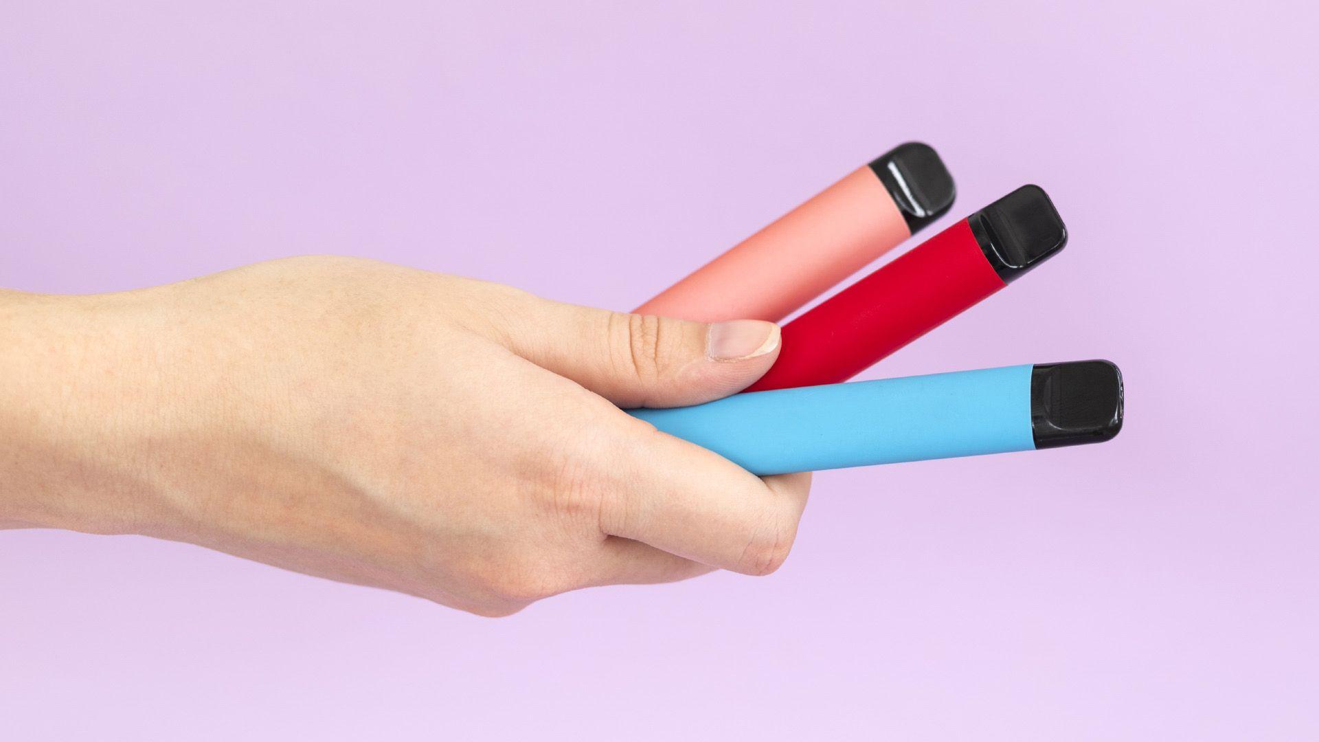 A white hand holding three disposable vapes. One is pink, one is red and one is blue