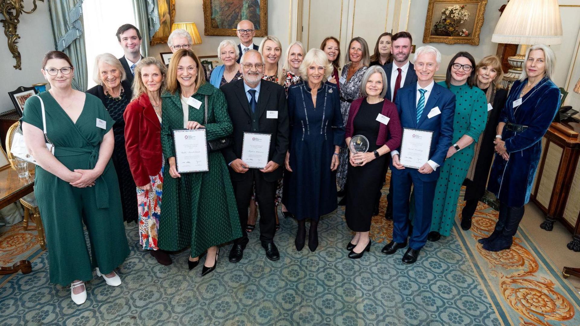 A group shot of doctors, volunteers and supporters at Clarence House in London. Everyone is dressed formerly and is smiling at the camera. They are standing on a faded blue carpet and several paintings are adorning the walls surrounding them.