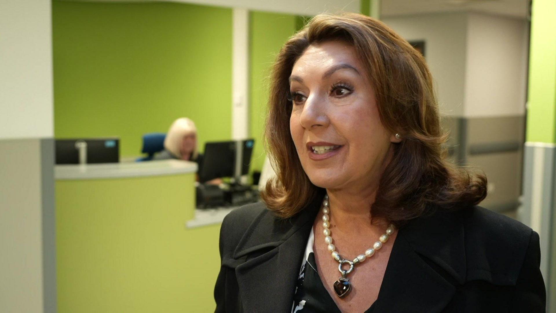 Jane McDonald, wearing a black jacket, standing in a hospital corridor with green walls.