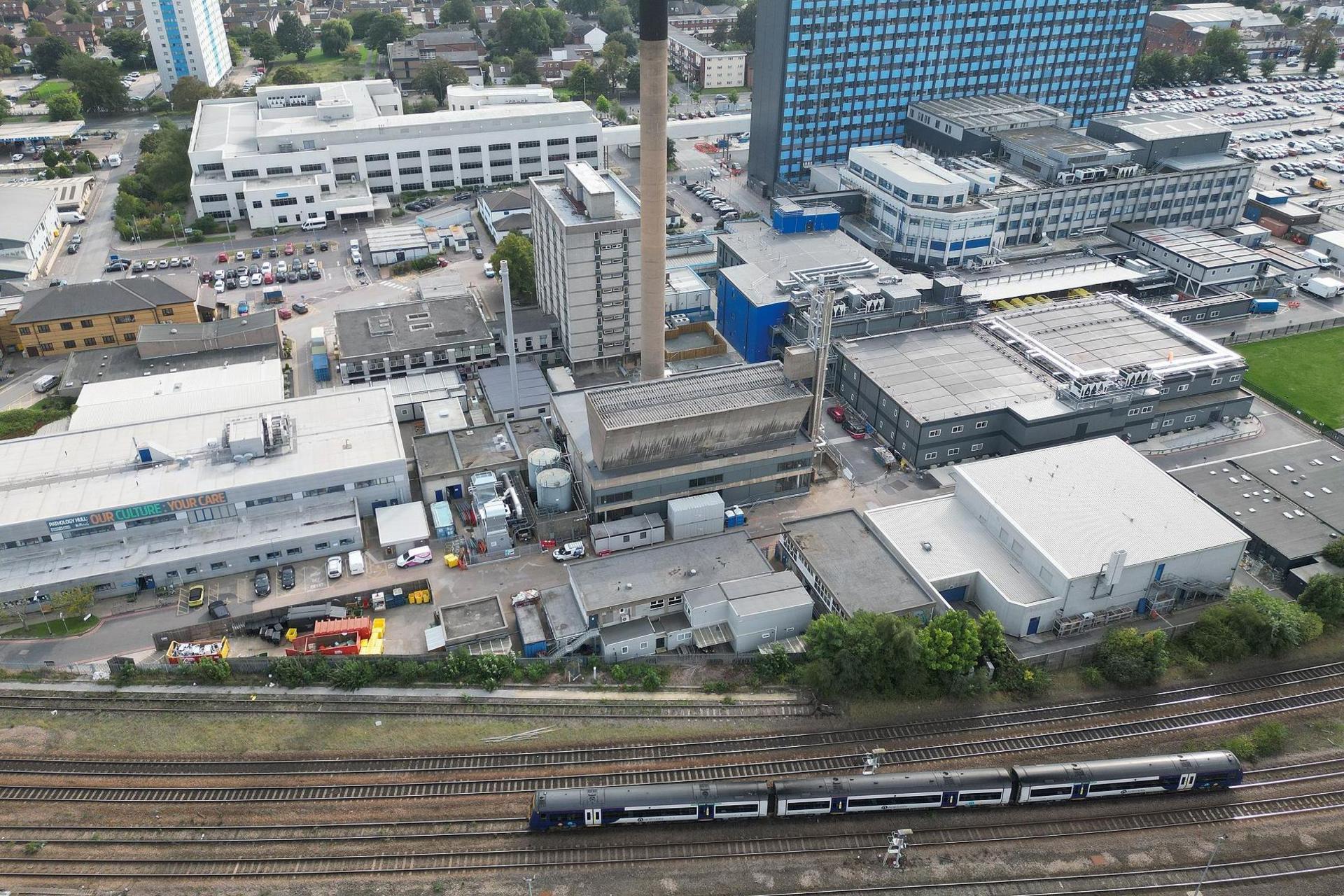 A train line with five tracks, one disused, runs by a large hospital estate. A black and white, three-carriage train is travelling by. The hospital estate is dominate by a large blue and grey tower block. Dozens of other smaller buildings surround it. A tall sand-coloured chimney rises in the centre of the view.