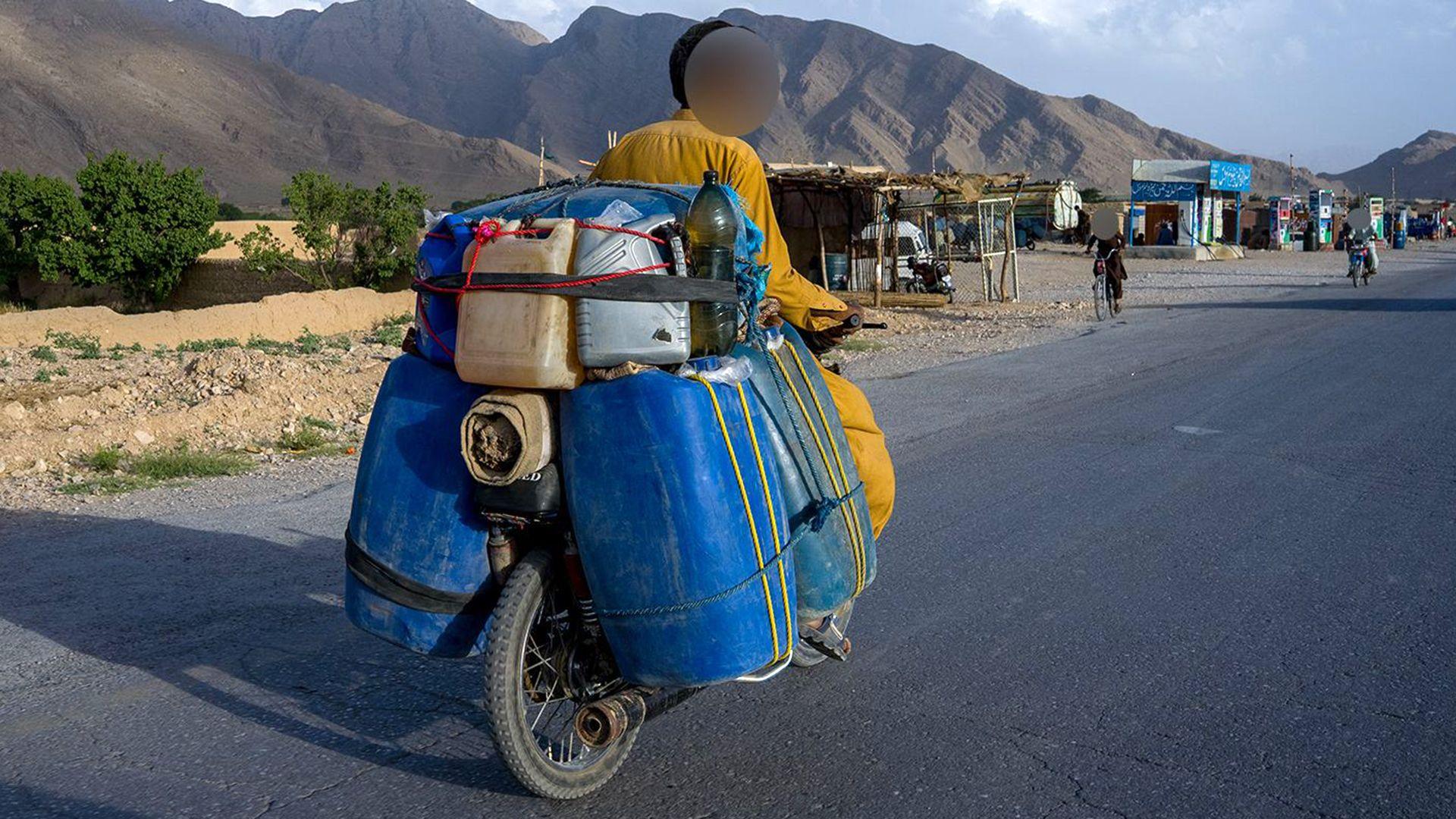 Mazaar is riding his small motorbike with canisters filled with fuel on the back of it. He's on a wide road in a semi-rural area with mountains in front of him and a few vehicles