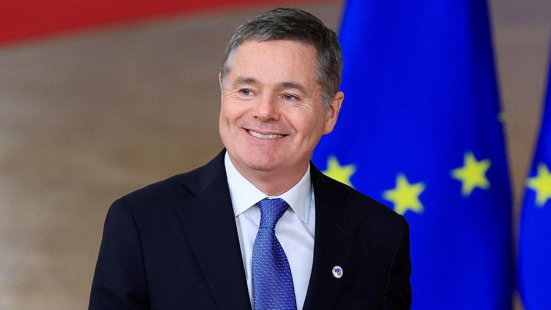 A close-up image of Paschal Donohoe attending a conference in Belgium. He is smiling at a camera. He is a middle-aged man with short, greying hair. He is wearing a dark suit jacket, a white collared shirt and a blue patterned tie.