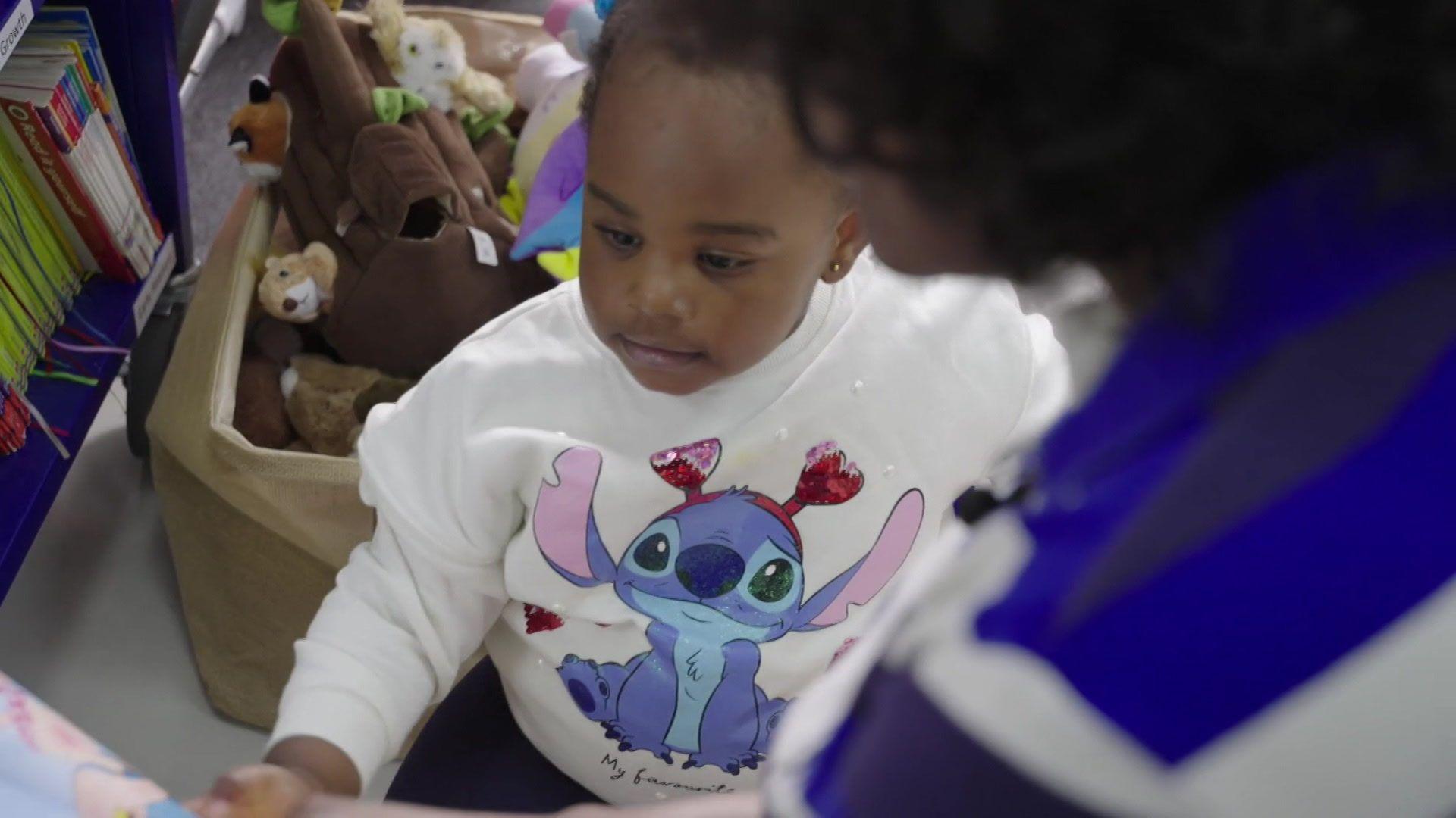 Emily wearing a white jumper with a big-eared purple cartoon character with red horns. Toys and books can be seen behind her