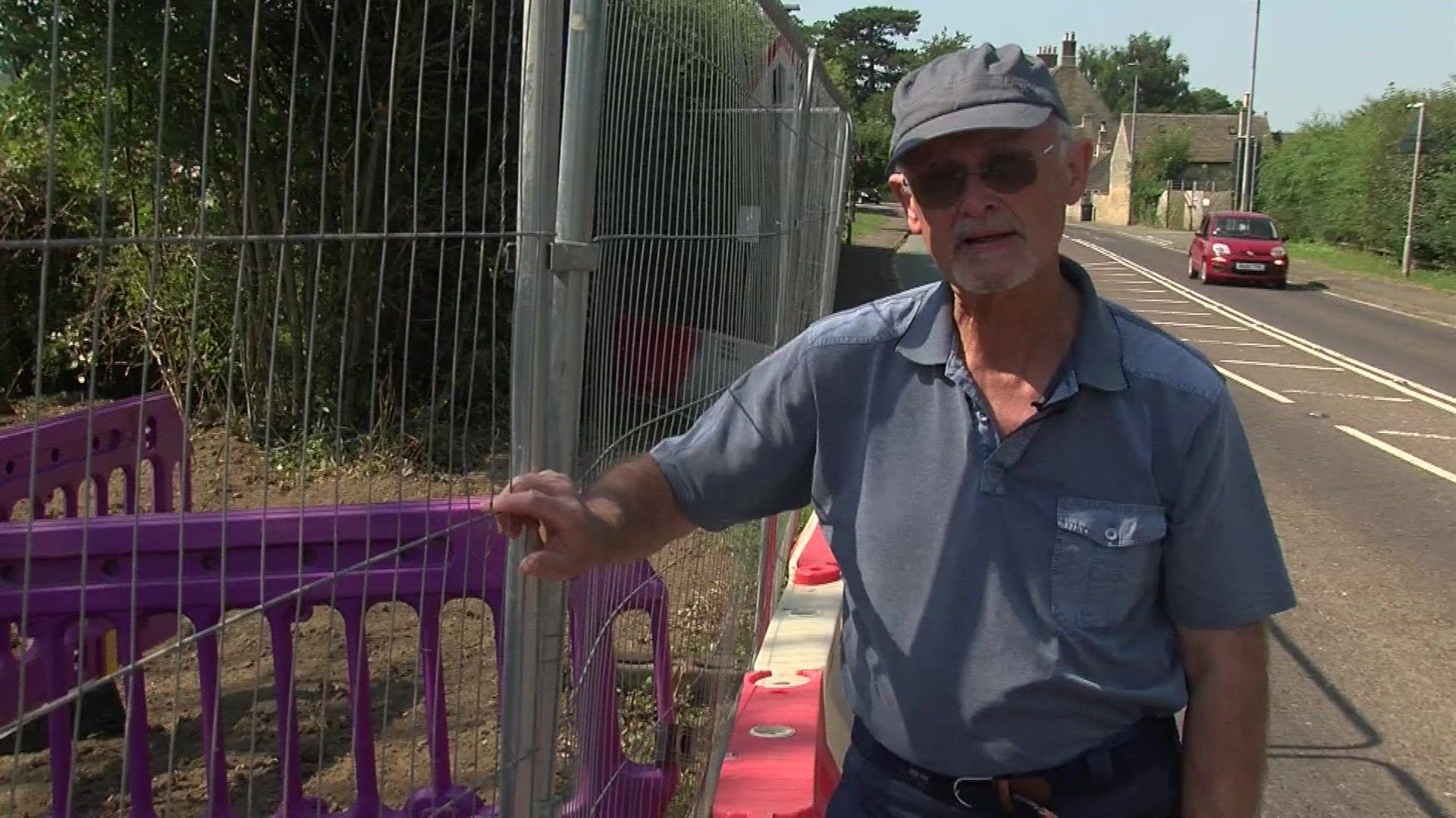 Tallington Parish Council chairman Philip Sagar stands next to a temporary metal fence, which surrounds purple safety barriers, by the side of a village road. He is wearing a blue polo shirt, a grey cap and dark glasses