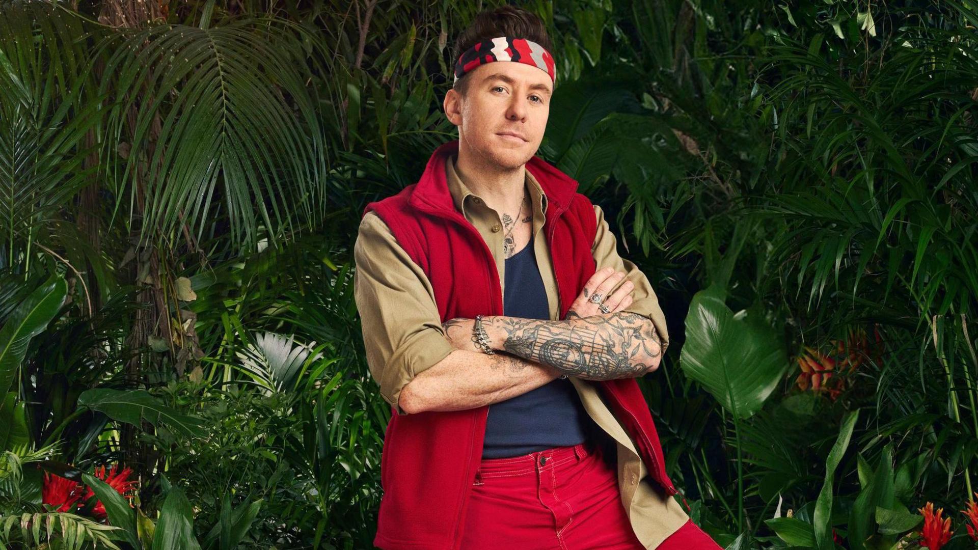 Danny Jones in front of jungle foliage folding his arms and smiling slightly into the camera