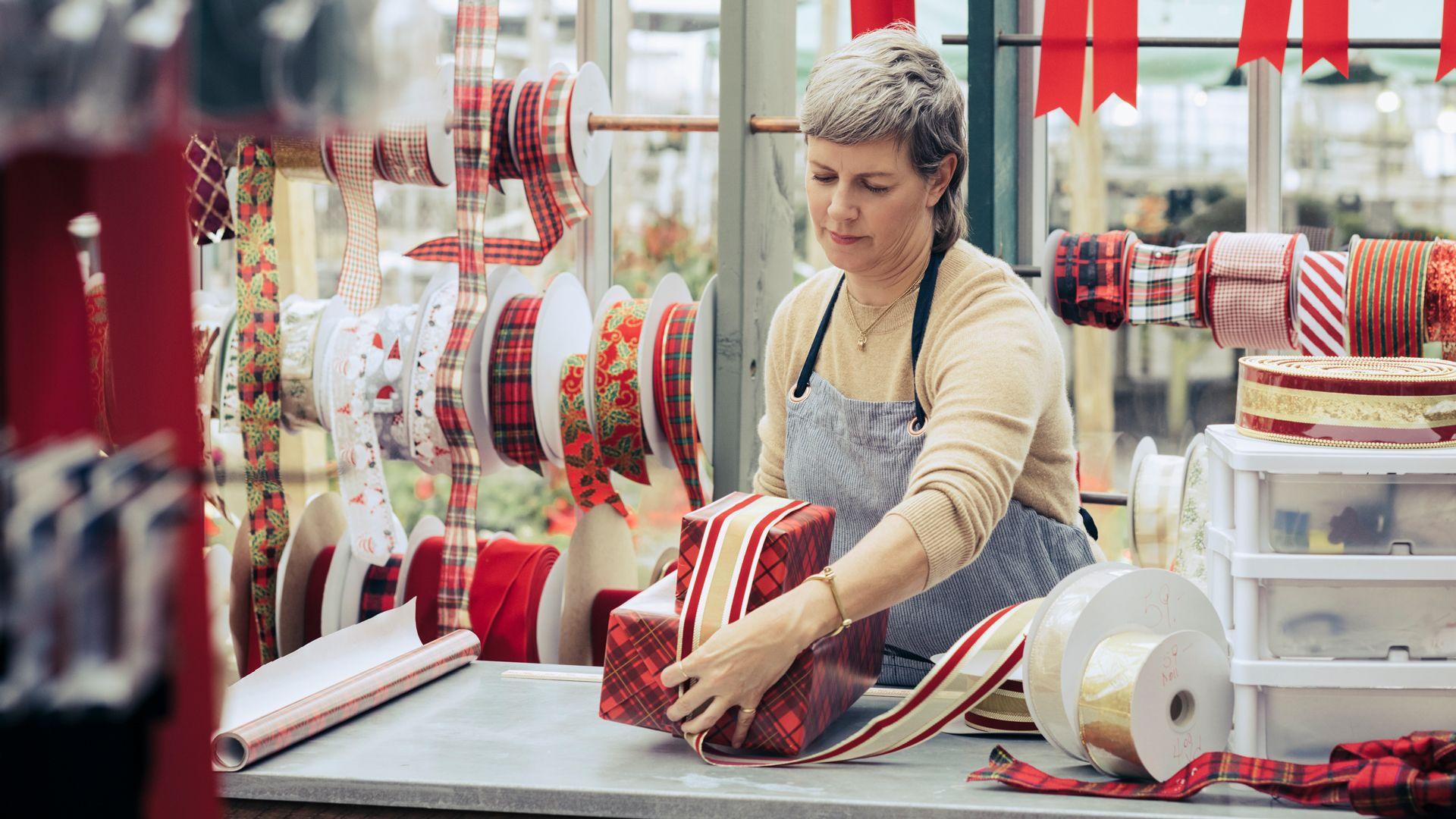A woman wearing an apron stands behind a counter wrapping Christmas presents with a thick red and gold ribbon. Reels of ribbons are seen behind her.