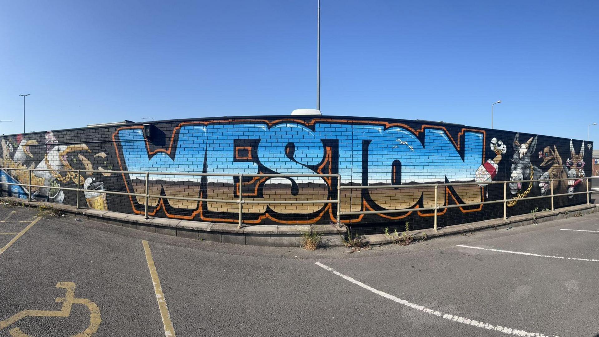 A wall is painted black with the word Weston painted in blue, white and brown which looks like a view of a beach