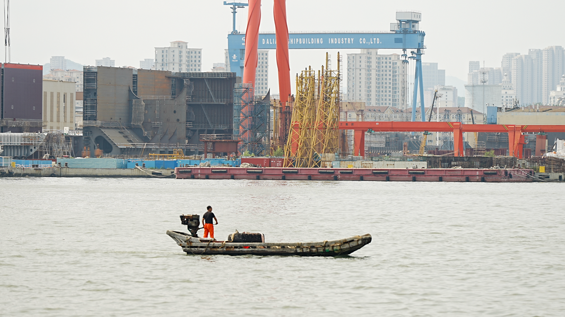 A man stands on a tiny vessel in the Dalian bay - in front of him is the shipyard with huge cranes and other equipment. A tall blue structure has the words Dalian Shipbuilding Industry Co. Ltd on it.