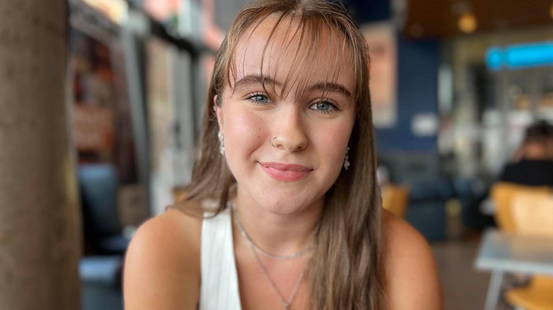 Millie Powell with long light brown hair, falling over her left shoulder, wearing  white top and slightly smiling with a nose ring, while sitting in a cafe