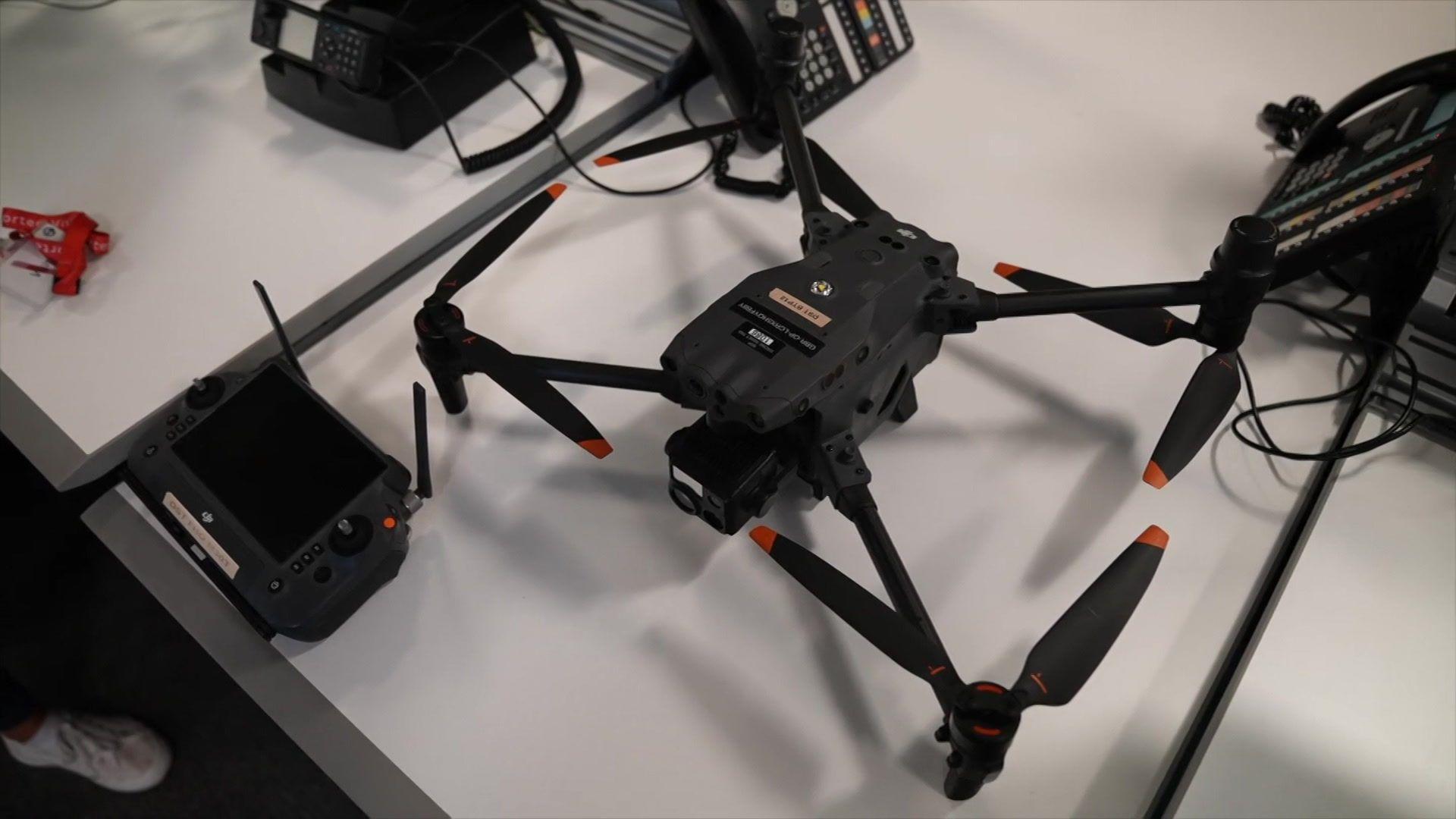 A sleek black drone with orange-tipped propellers sits on a white desk next to its remote control. The desk also has office items on it like telephones and cables.