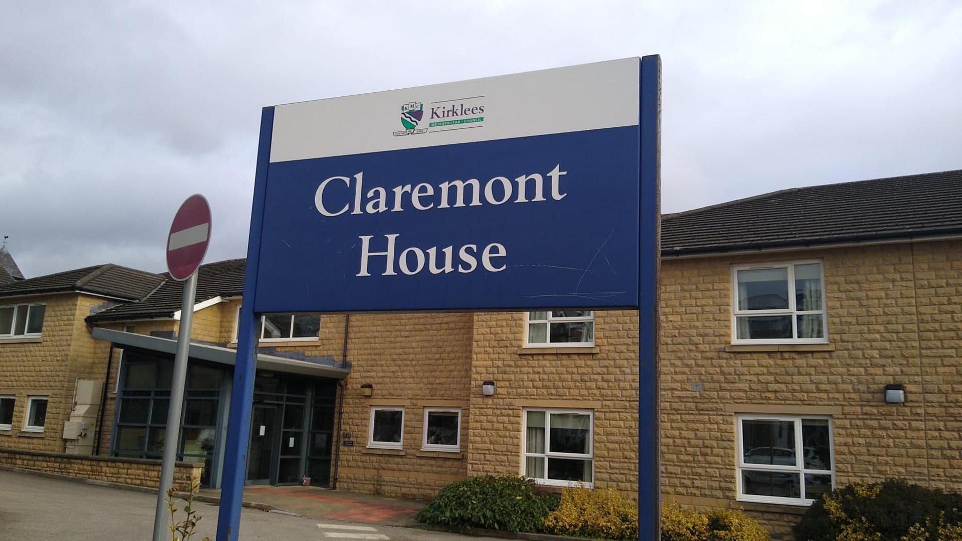 A two storey building with sand coloured bricks and windows with white frames. 
In the foreground is a blue sign with 'Claremont House' the name of the dementia home on. 