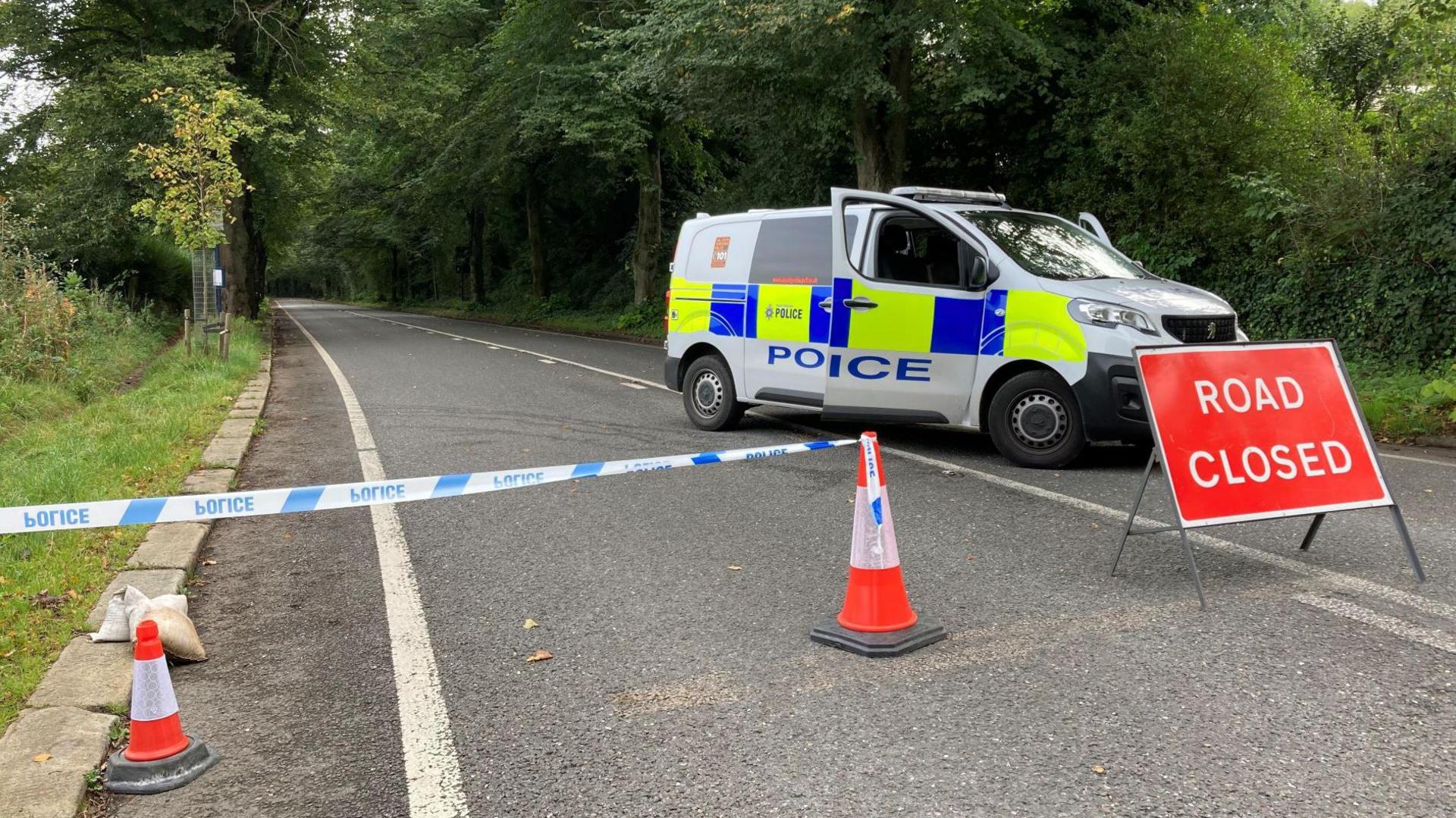 The image shows a police cordon extending to the middle of the road. A van is parked on the right, blocking the way, with a red "Road closed" sign in front of it.