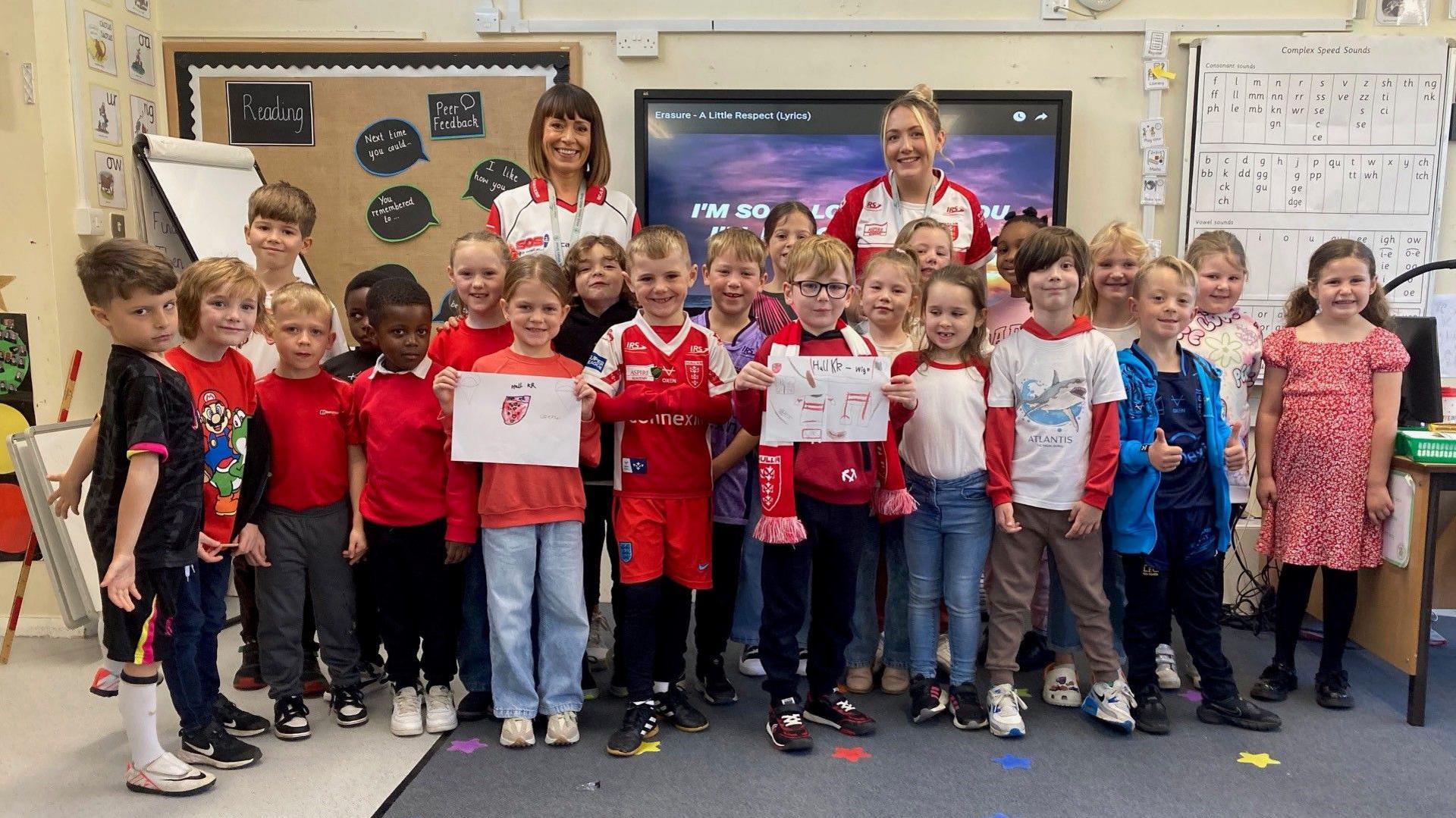 A group of children stand in a classroom and hold up drawings as part of their red and white day.