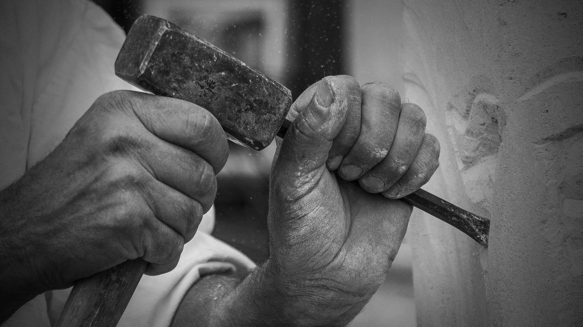 A stock black and white close-up image of the hands of a stonemason working a block of stone with a hammer and chisel. 