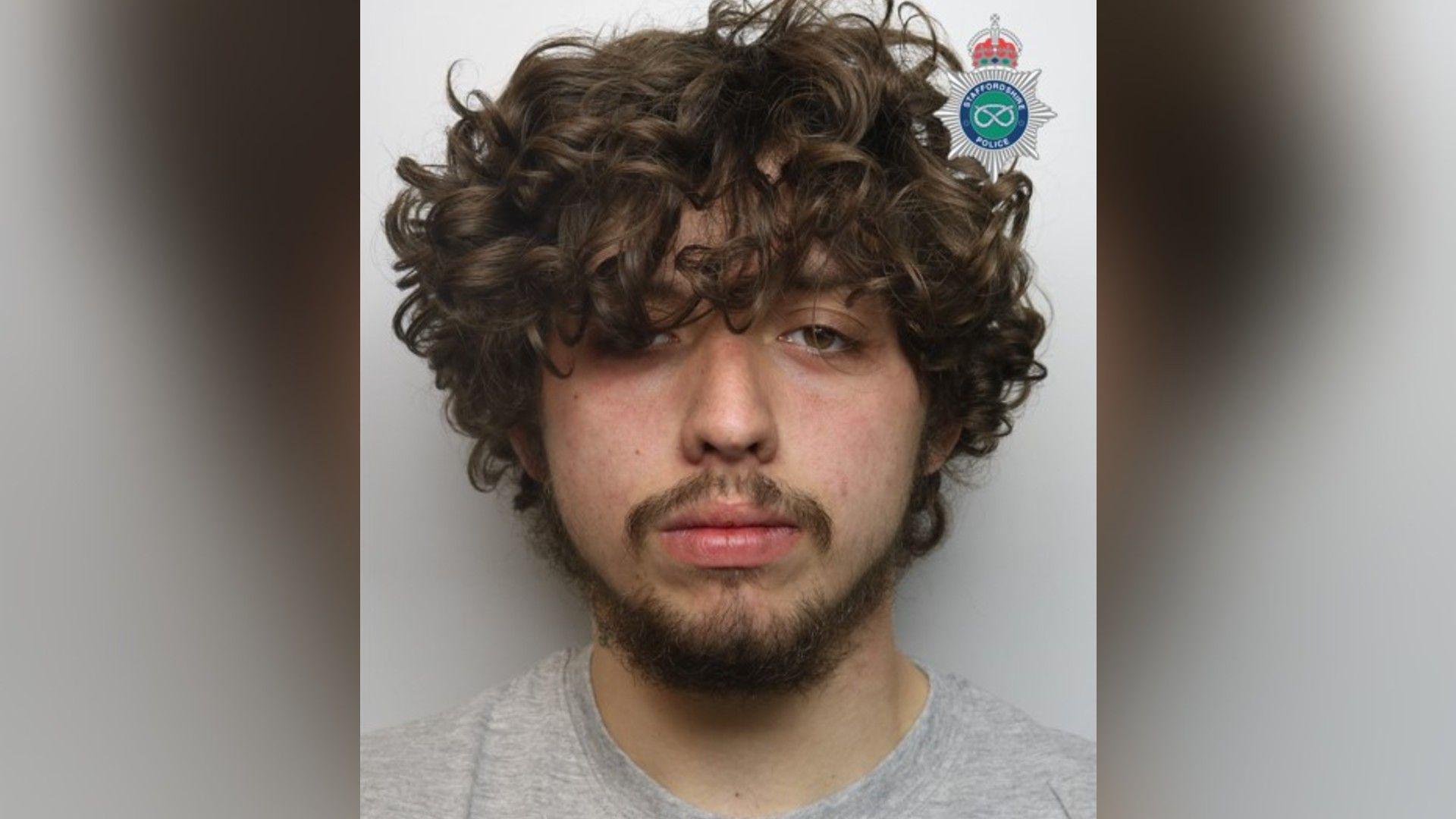 A mugshot of a man with brown curly hair, which is partially covering his eyes, with a black beard and a grey shirt.
