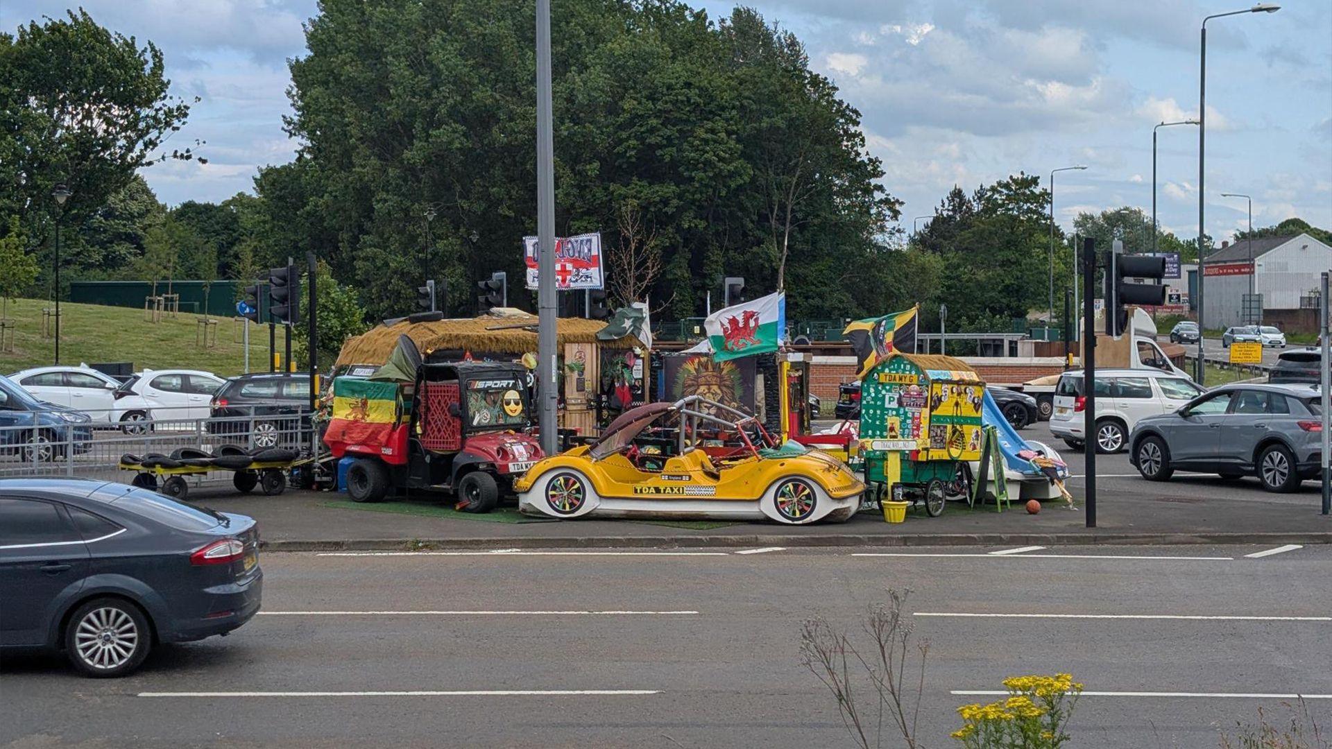 A bright yellow car, caravan and other brightly-coloured pieces of equipment on an island off a busy main road
