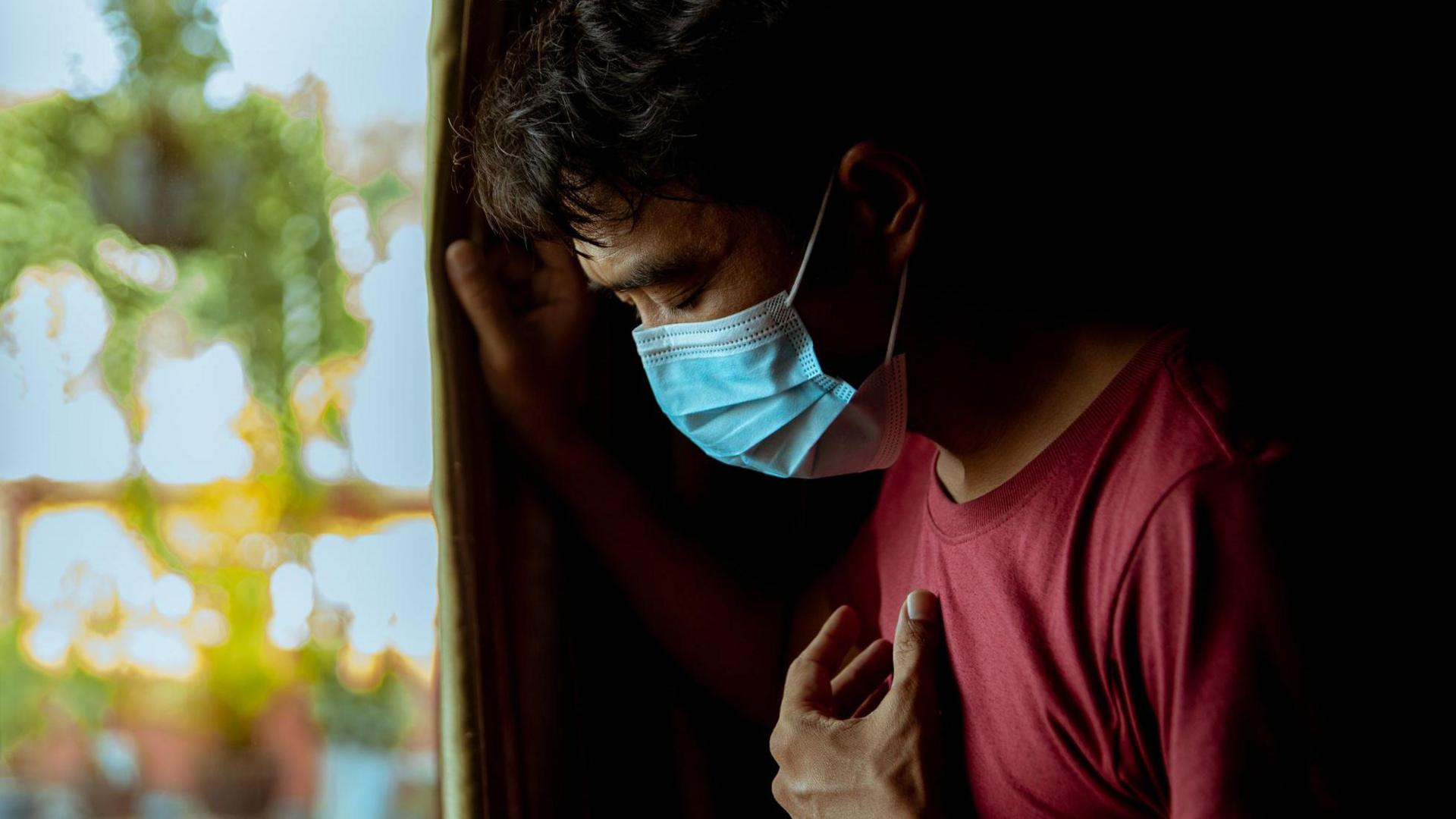 An asian man of unknown age stands by a window in a red tshirt and wearing a blue face mask. HIs head is bowed and his eyes are closed as if sad or contemplative and he holds a hand to his chest. 
