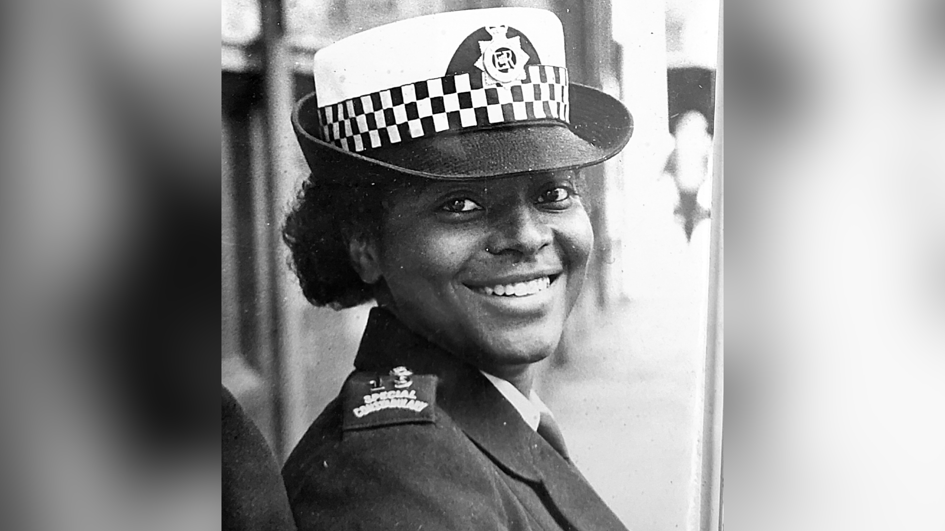 A black and white photo of Beverly Douglas. She is wearing police uniform, including a black and white hat and a black jacket with special constable on the shoulder pads. She is looking at the camera and smiling.